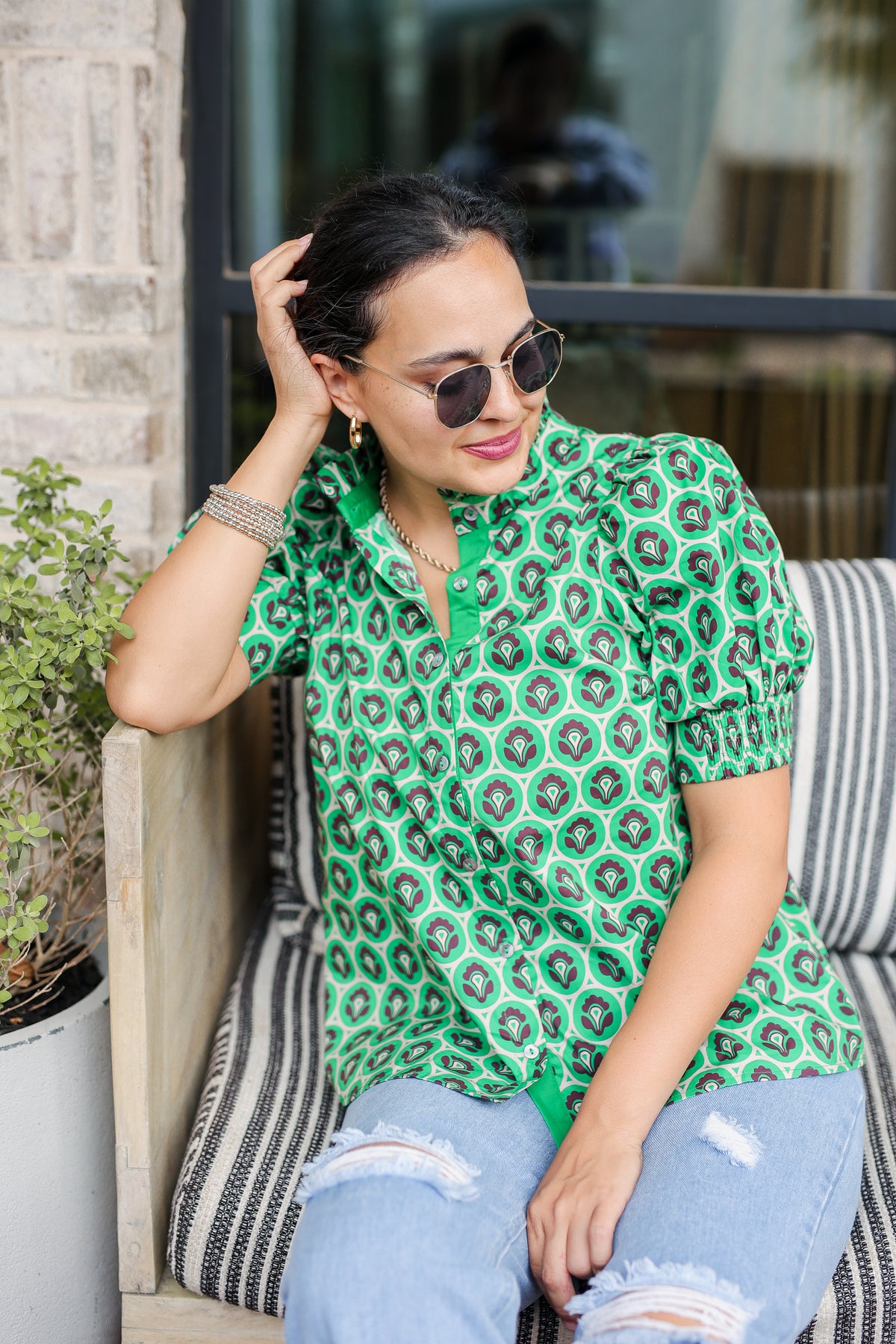 Woman wearing a green patterned shirt and sunglasses, sitting on a bench.