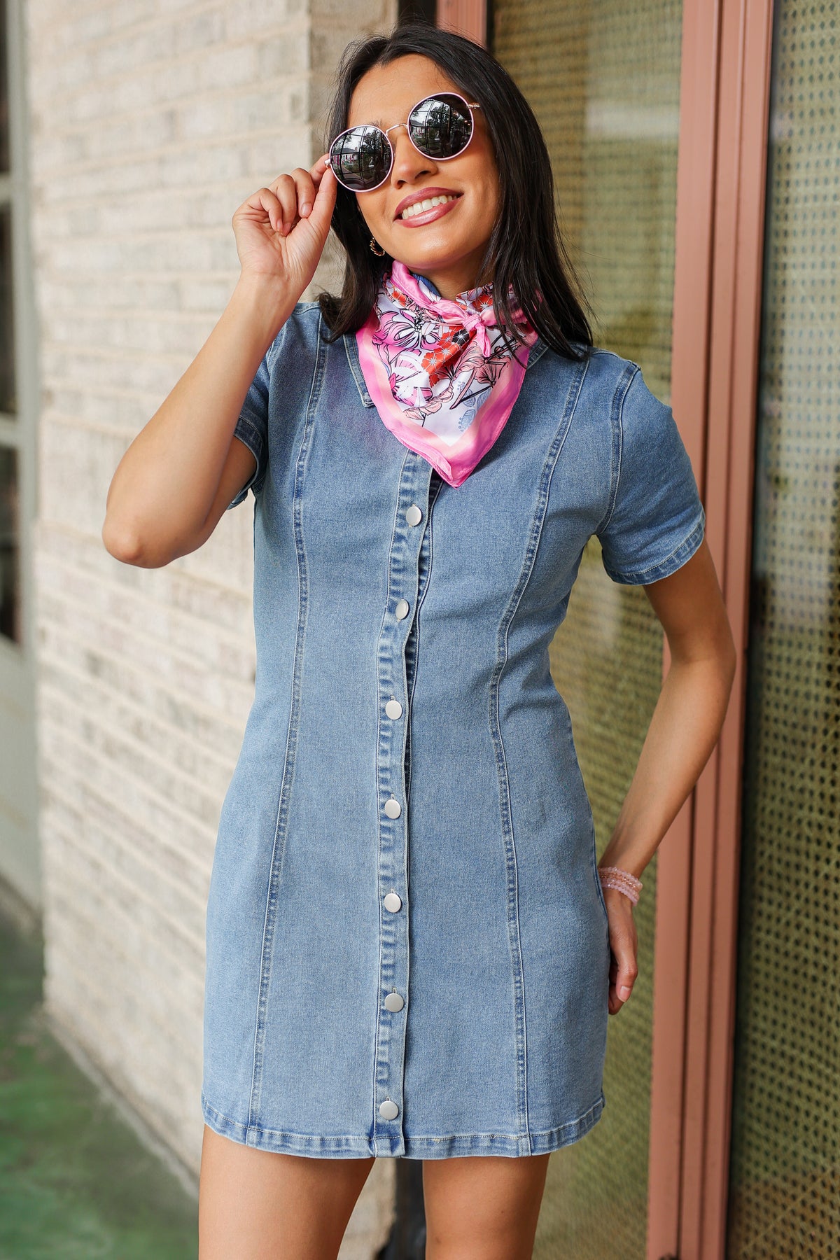 Woman wearing a denim dress with a pink scarf, standing against a textured wall.