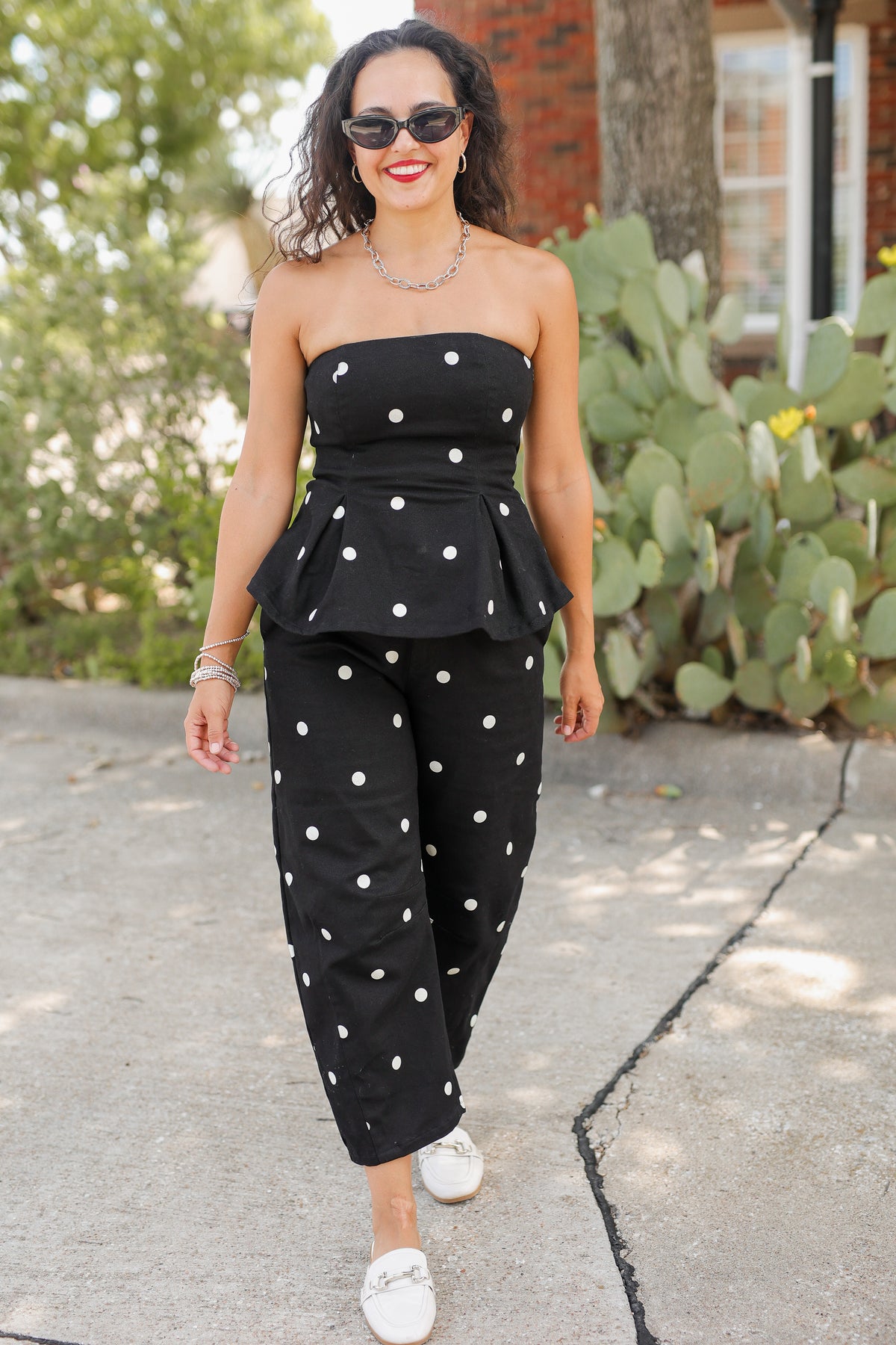 A woman wearing a black strapless peplum top and matching wide-leg pants with white polka dots, walking outdoors on a paved path with cacti and a brick building in the background.