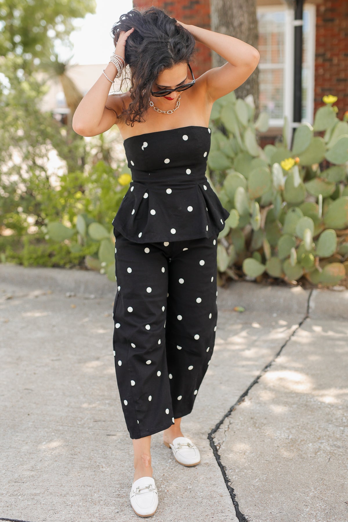 A woman wearing a black strapless jumpsuit with white polka dots and white slip-on shoes, standing outdoors on a concrete path with cacti and greenery in the background.