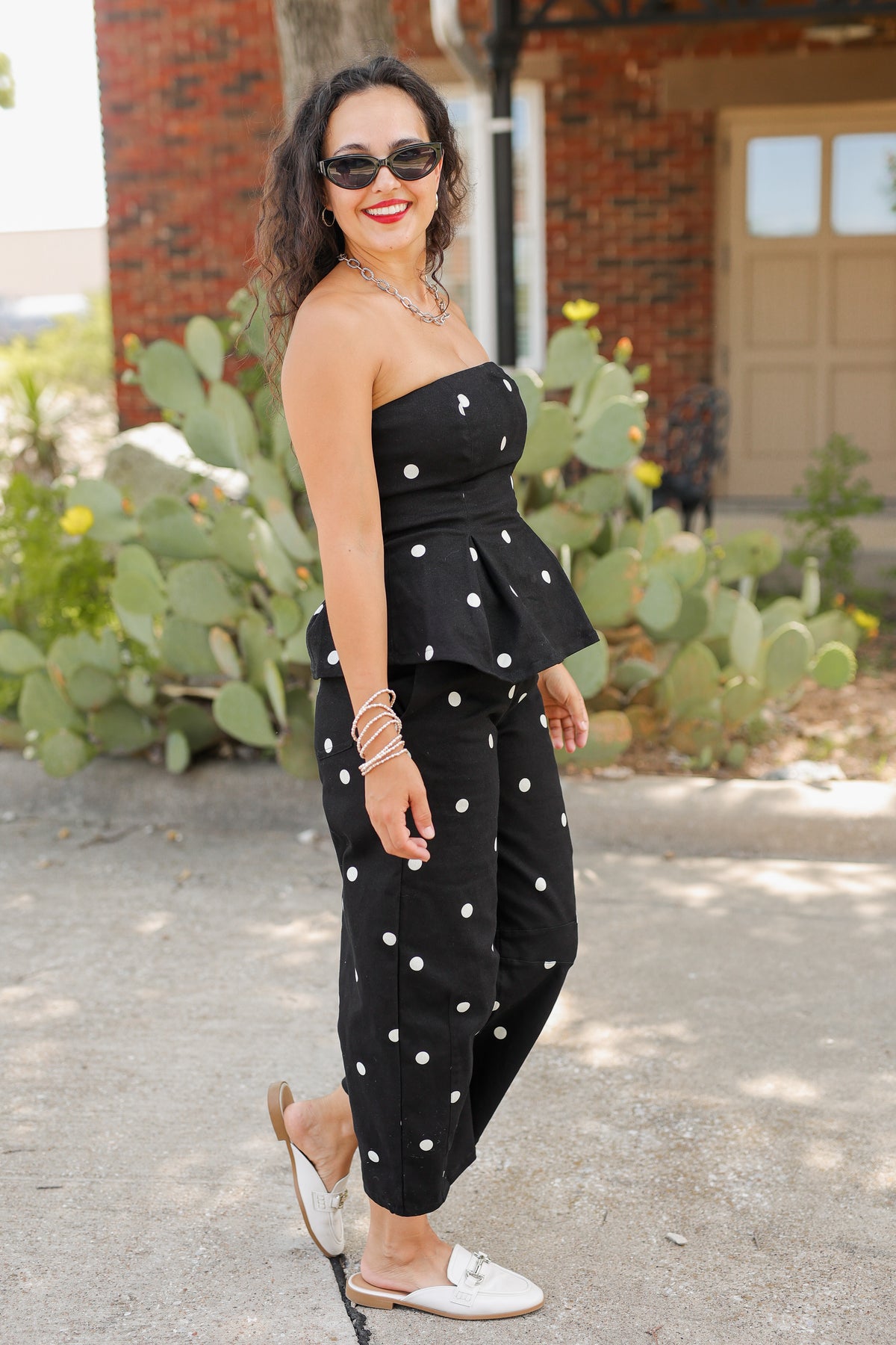 A woman wearing a black strapless jumpsuit with white polka dots, styled with white slide sandals, sunglasses, and bracelets, standing outdoors in front of a brick building and cacti.
