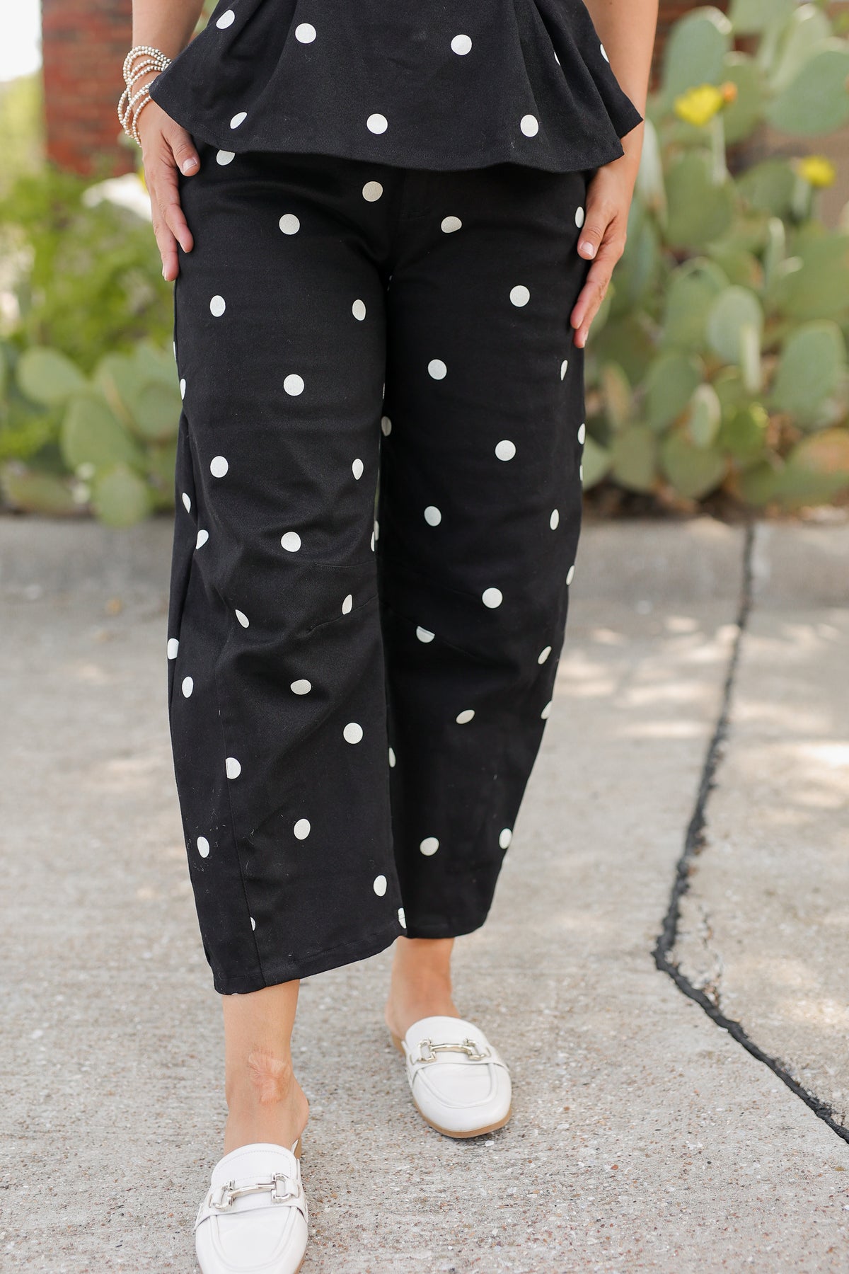 A person wearing black cropped pants with white polka dots, paired with a matching black top and white loafers, standing outdoors on a paved walkway with cacti in the background.