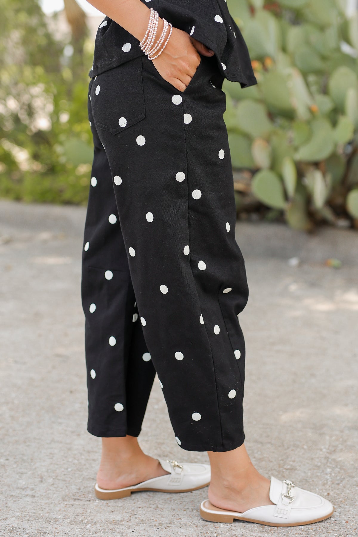 Black wide-leg cropped pants with white polka dots, worn by a model standing outdoors on a paved surface with greenery in the background.