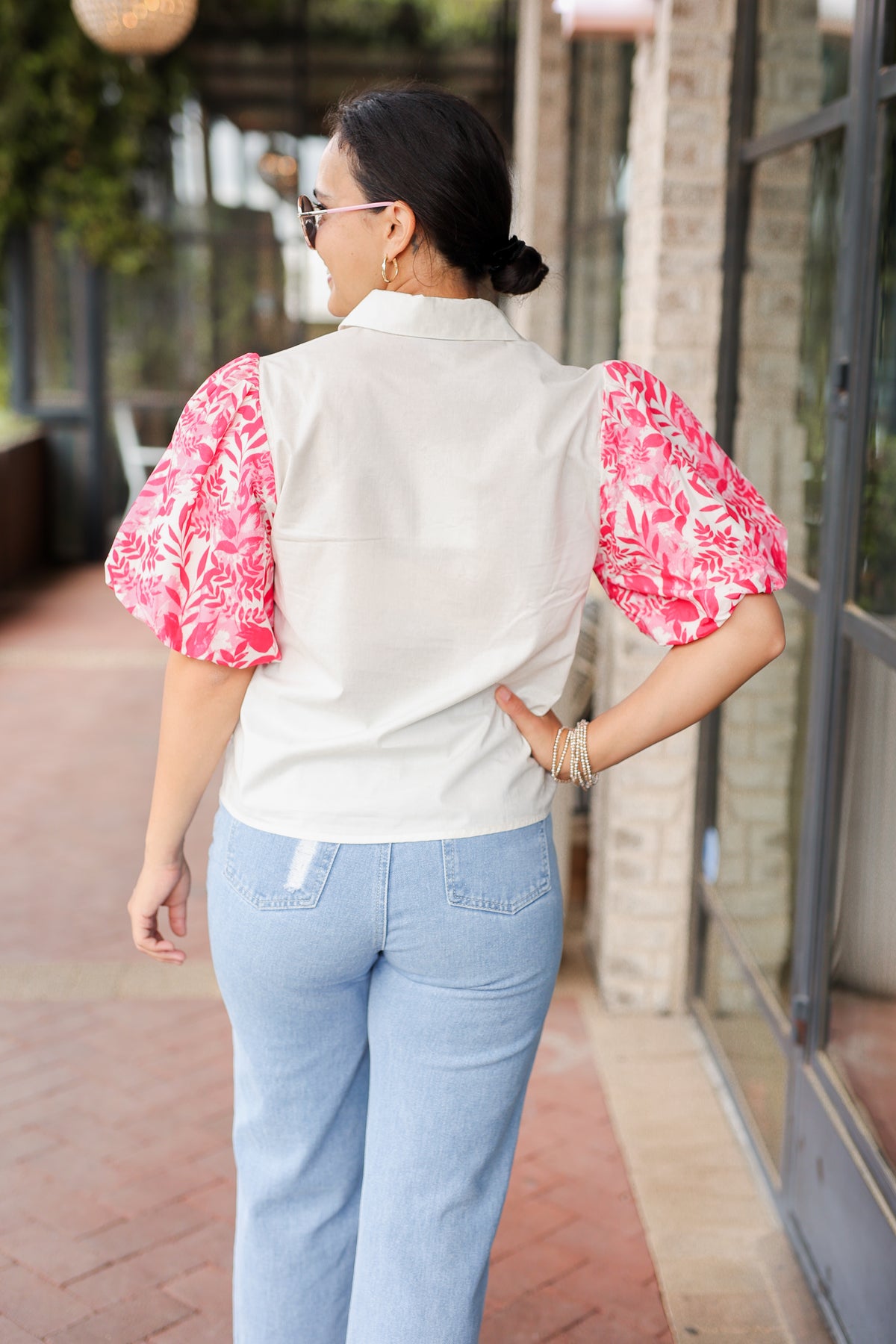 Woman wearing a white blouse with pink floral sleeves and light blue jeans, standing outdoors.