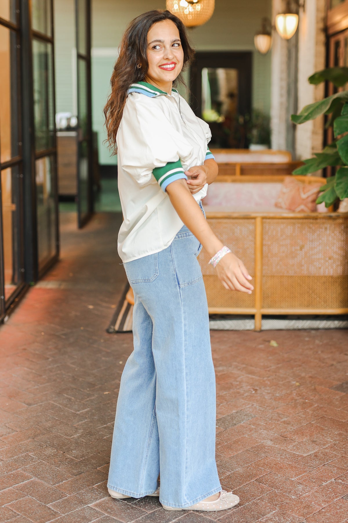 Woman in a white blouse and light blue jeans standing in an indoor setting with plants and furniture.