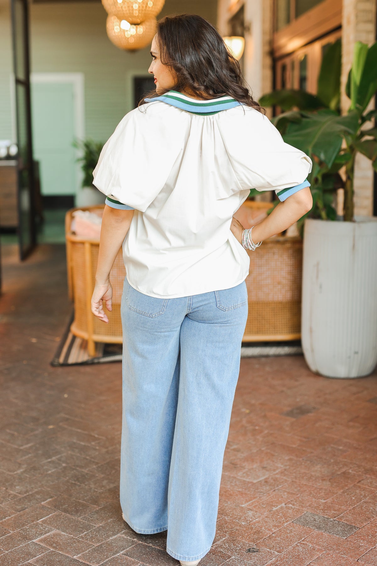 Woman wearing a white blouse and light blue jeans standing outdoors with plants and furniture in the background.