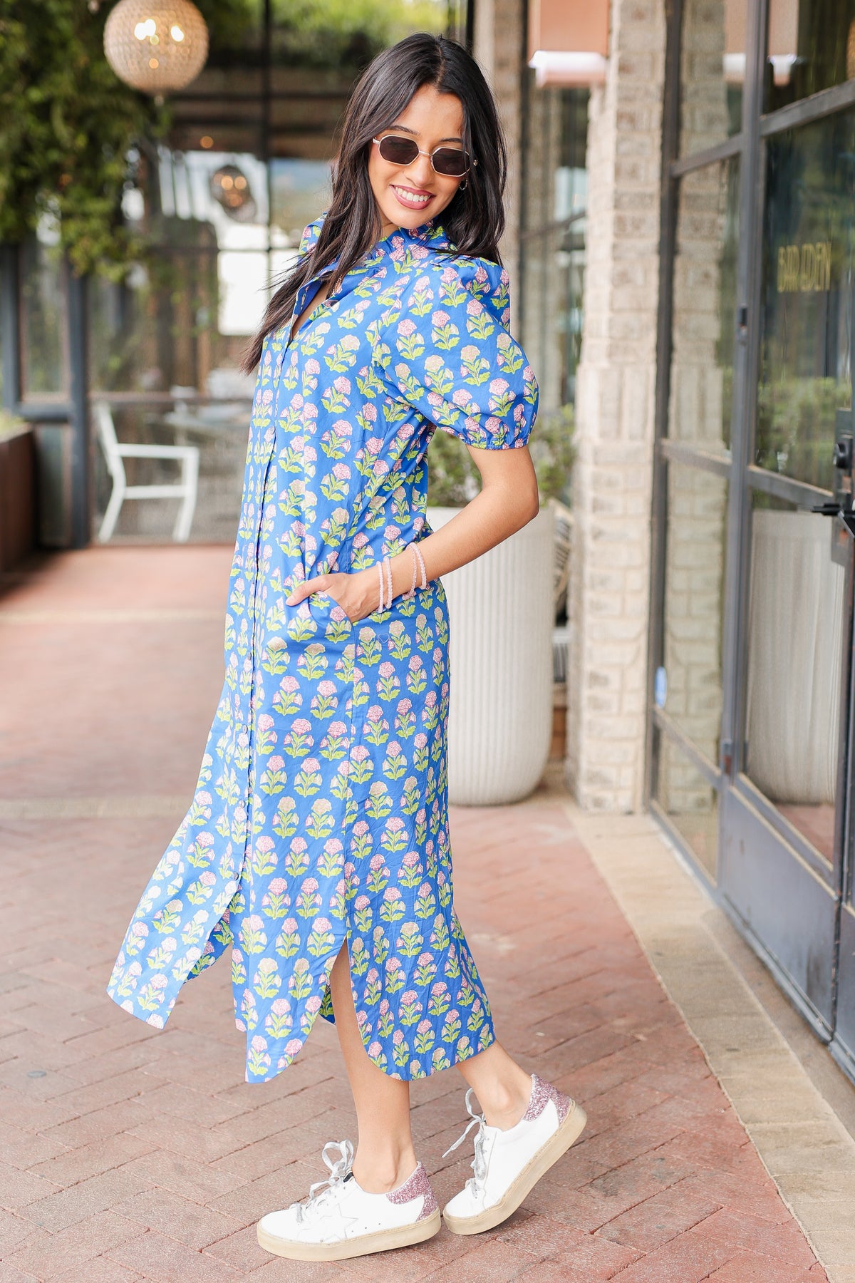 Woman in a blue floral dress standing outdoors near a building entrance.