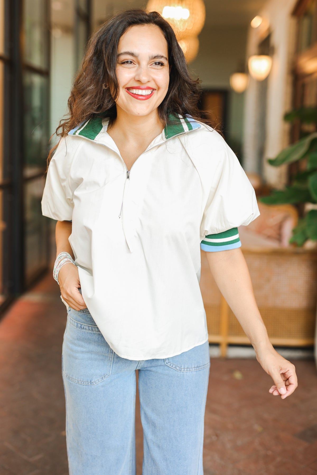 Woman wearing a white blouse with green accents and blue jeans, standing in an indoor setting.
