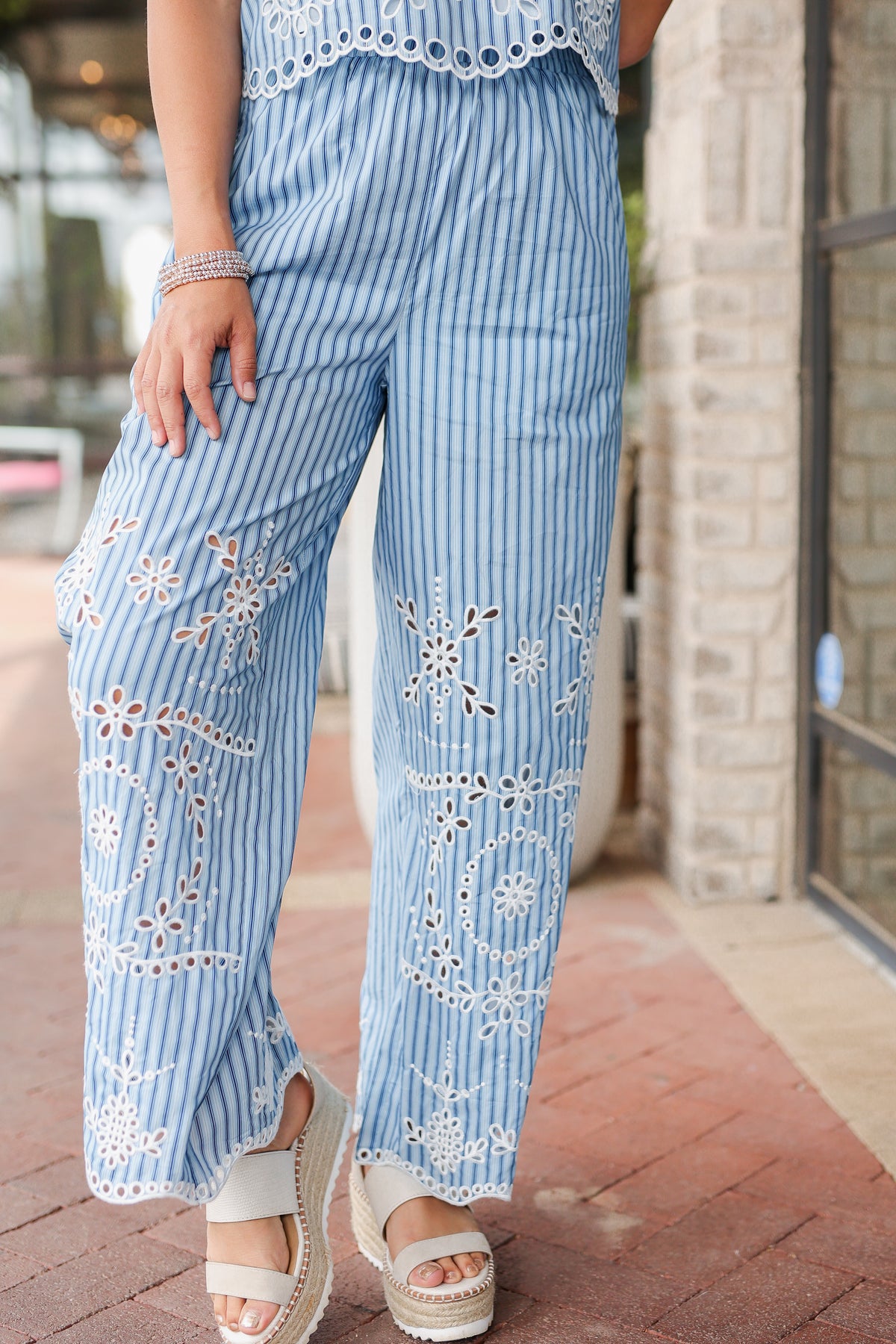 Woman wearing blue striped pants with white embroidery and sandals on a sidewalk.
