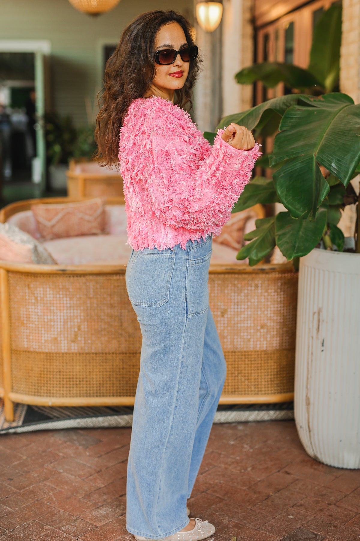 Woman wearing a pink textured sweater and blue jeans standing in an outdoor setting with plants and furniture.