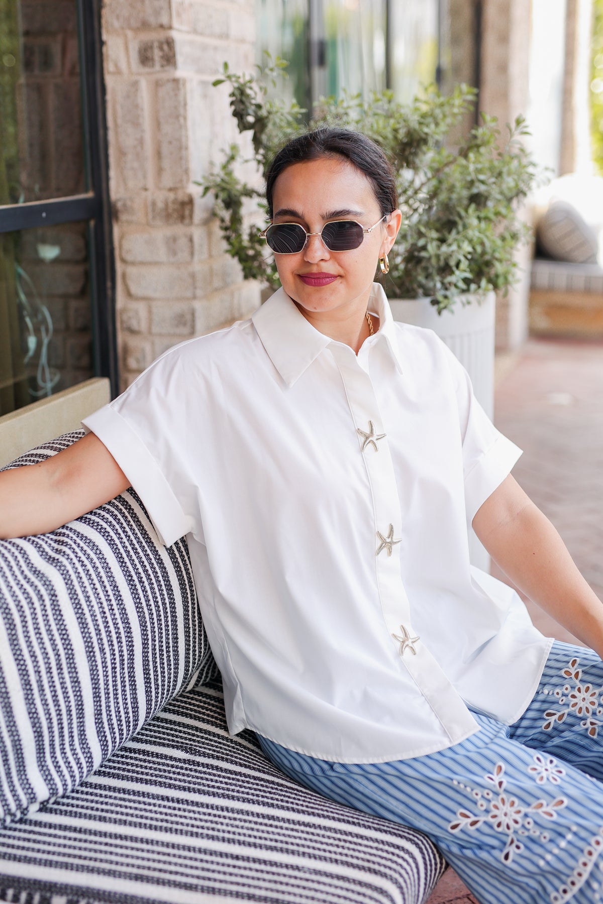 Woman wearing sunglasses and a white shirt with a pattern, sitting on a striped couch.