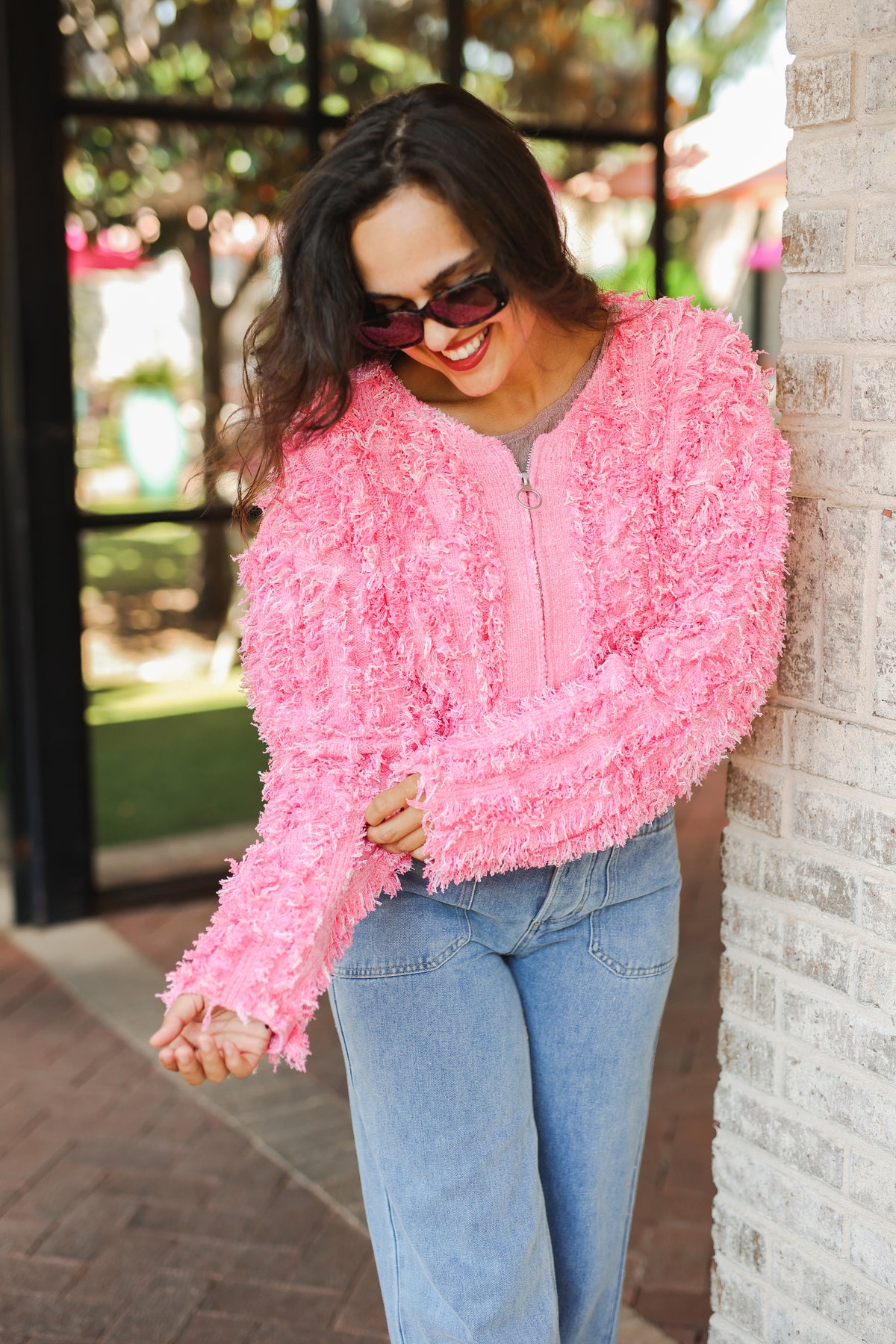 Woman wearing a pink textured sweater and sunglasses, leaning against a brick wall.