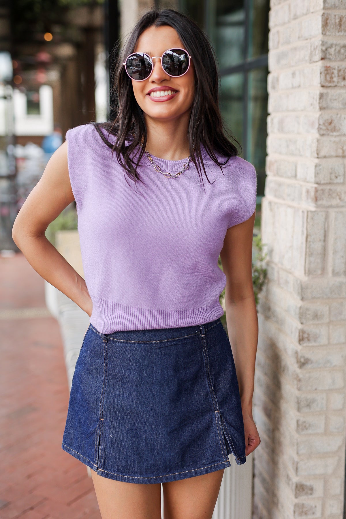 Woman wearing a purple sleeveless top and blue denim skirt standing against a brick wall.