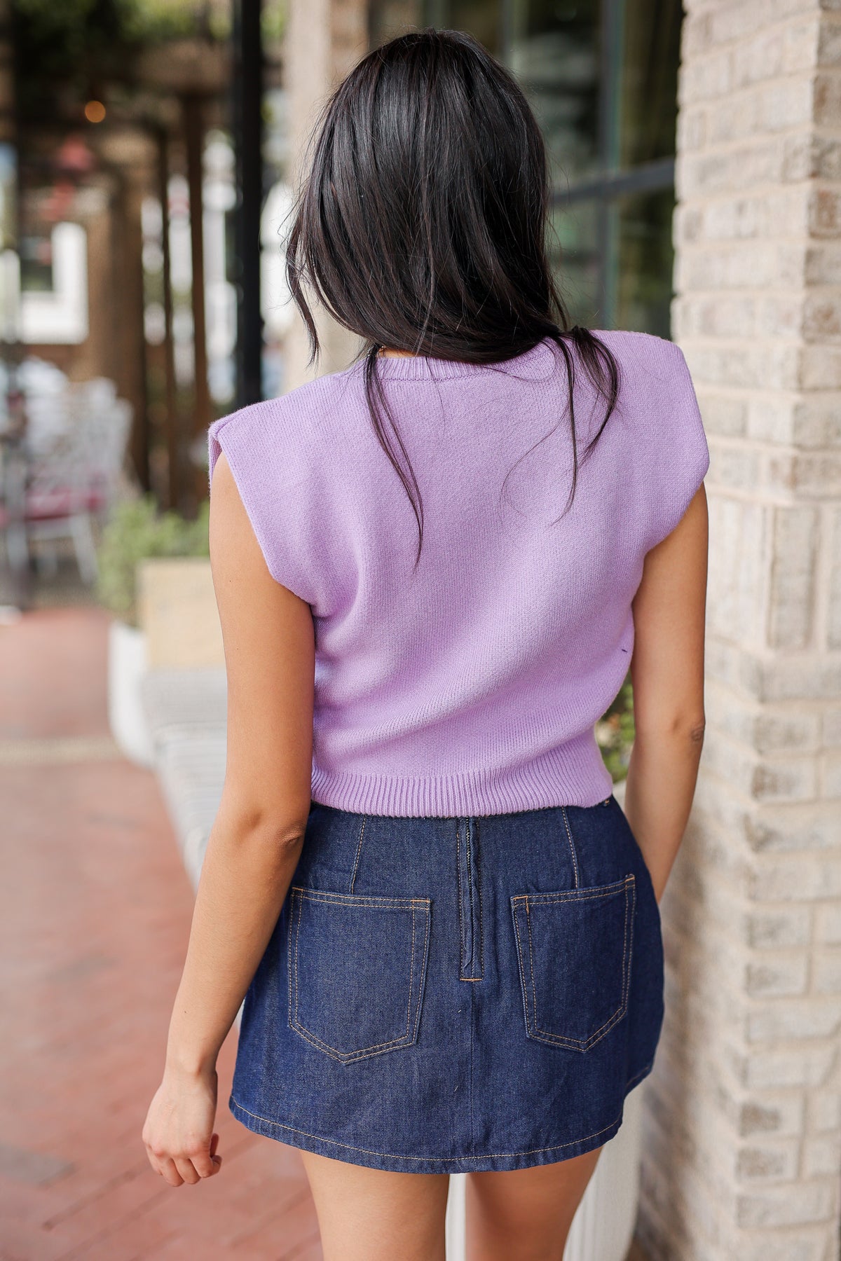 Person wearing a purple sleeveless top and denim skirt standing against a brick wall.