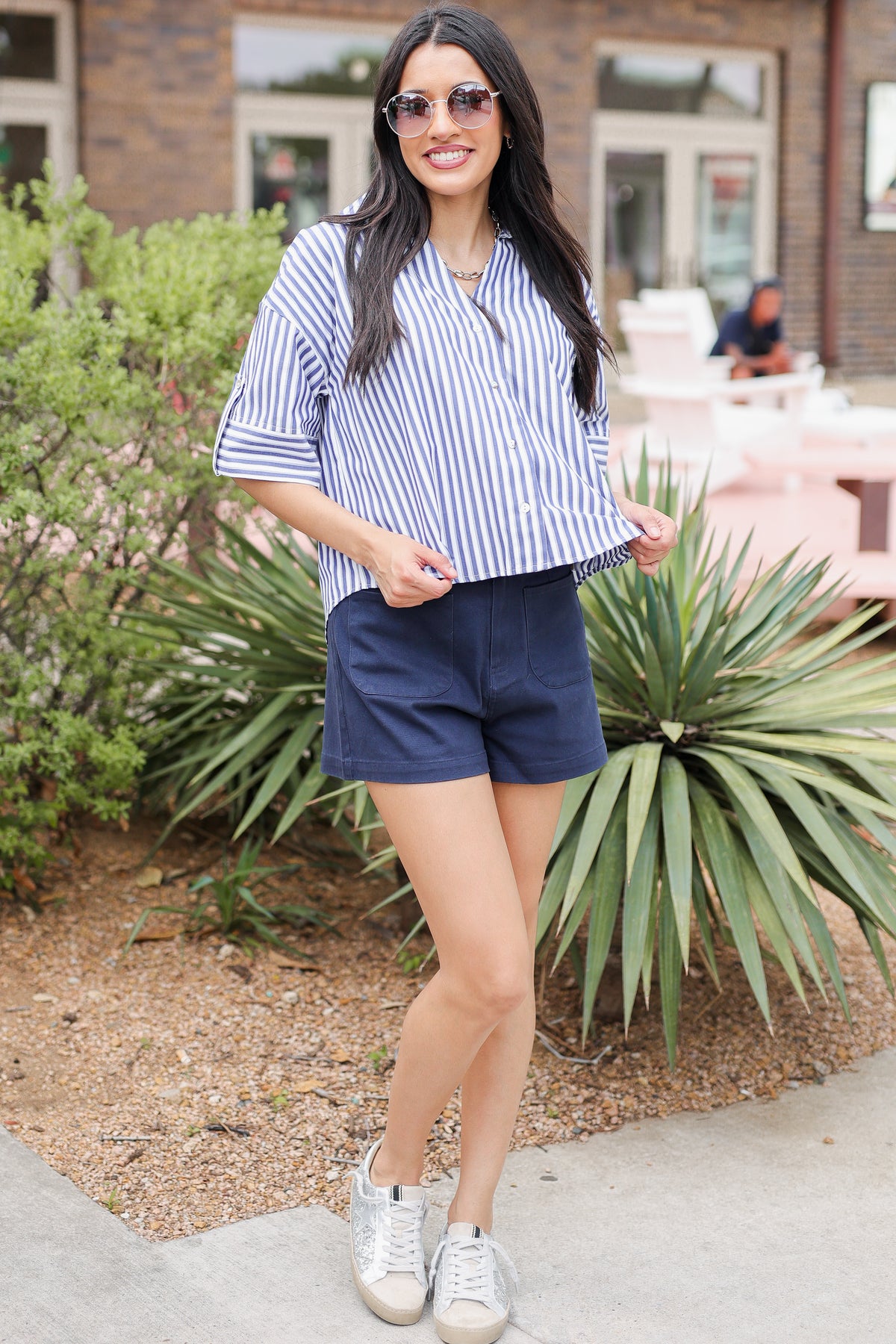 Woman in a striped shirt and shorts standing outdoors with plants and a building in the background