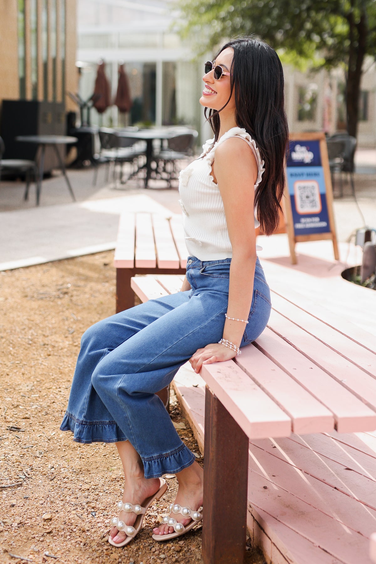 Woman sitting on a pink bench wearing a white top and blue jeans, with a blurred outdoor background.