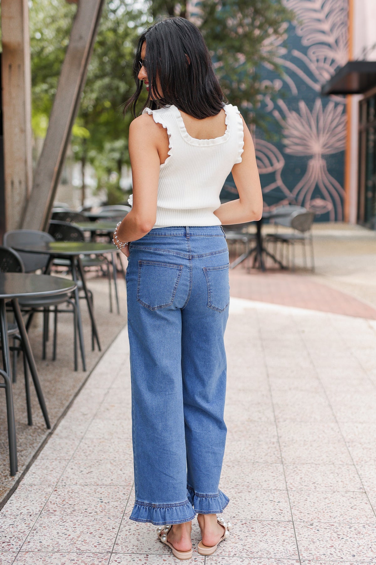 Woman in a white top and blue jeans standing on a sidewalk with outdoor furniture in the background.