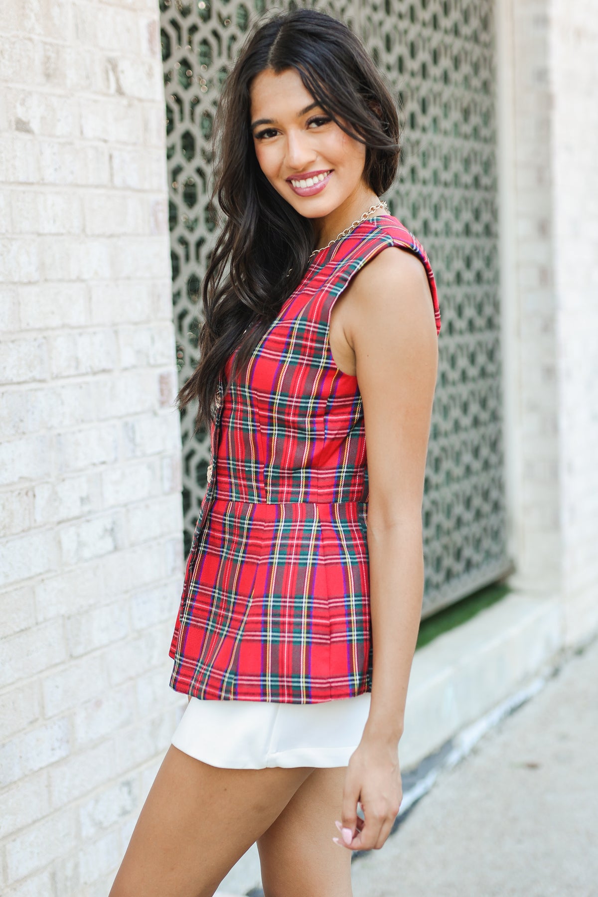 Woman wearing a red plaid sleeveless top and white shorts standing against a textured wall.