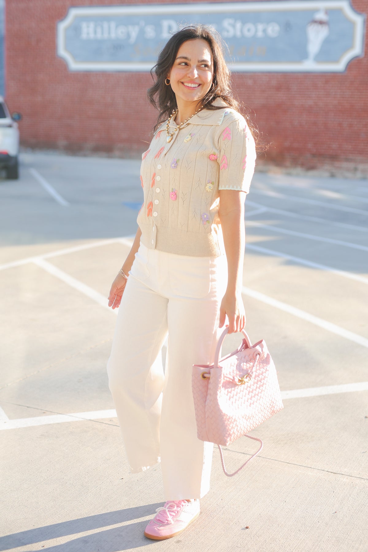 Woman in a light-colored outfit with a pink handbag standing in a parking lot.