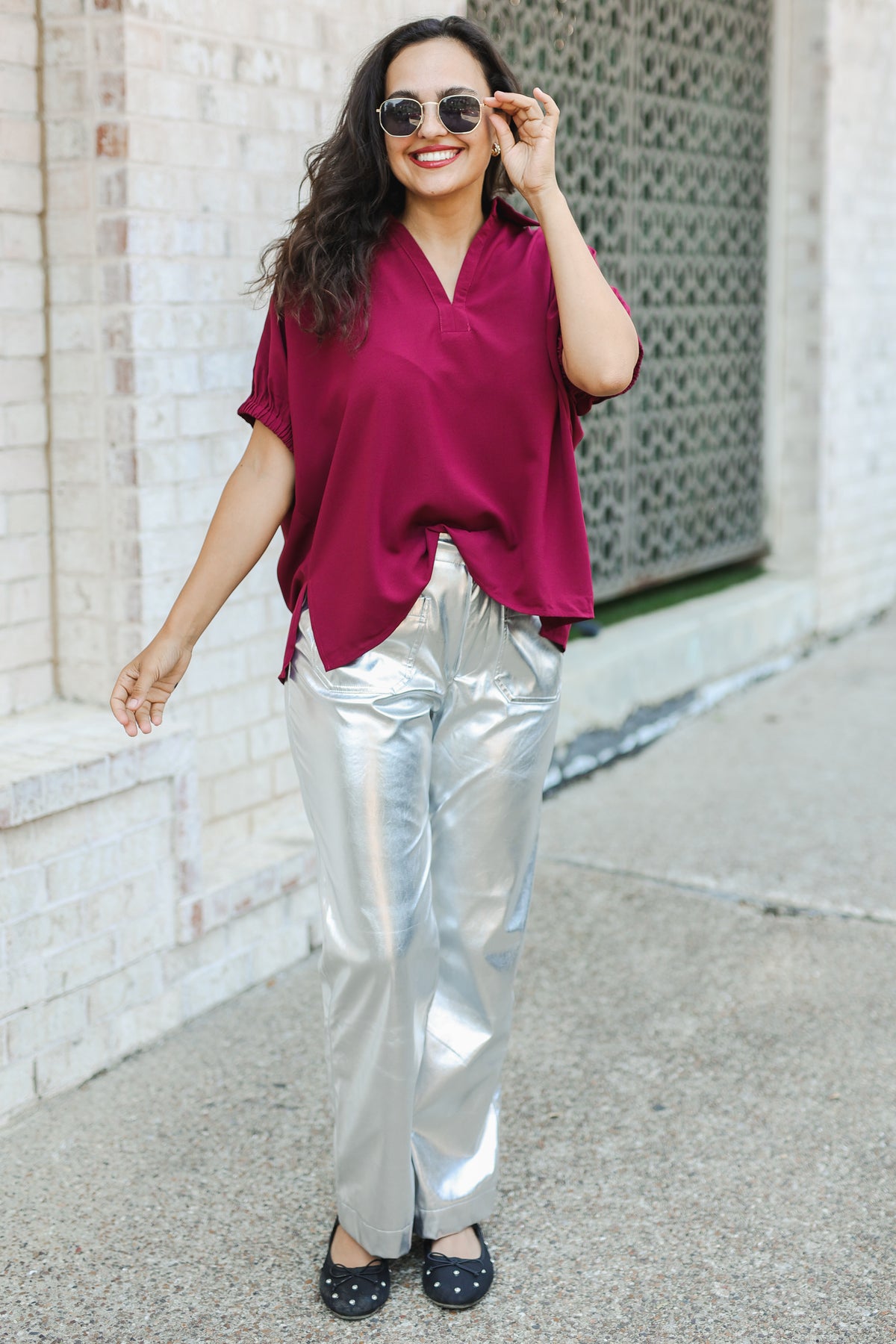 Woman wearing a red top and silver pants standing against a white brick wall.
