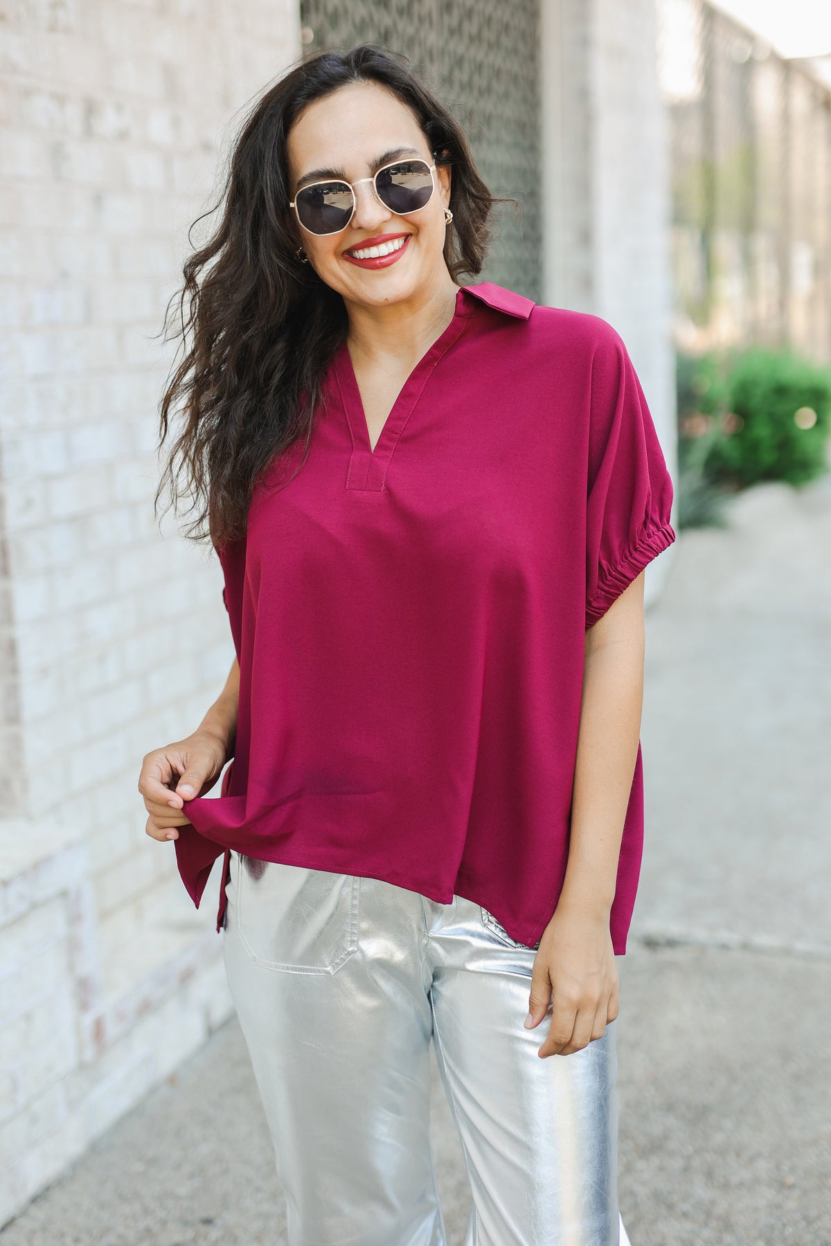 Woman wearing a magenta shirt and silver pants standing against a light-colored wall.