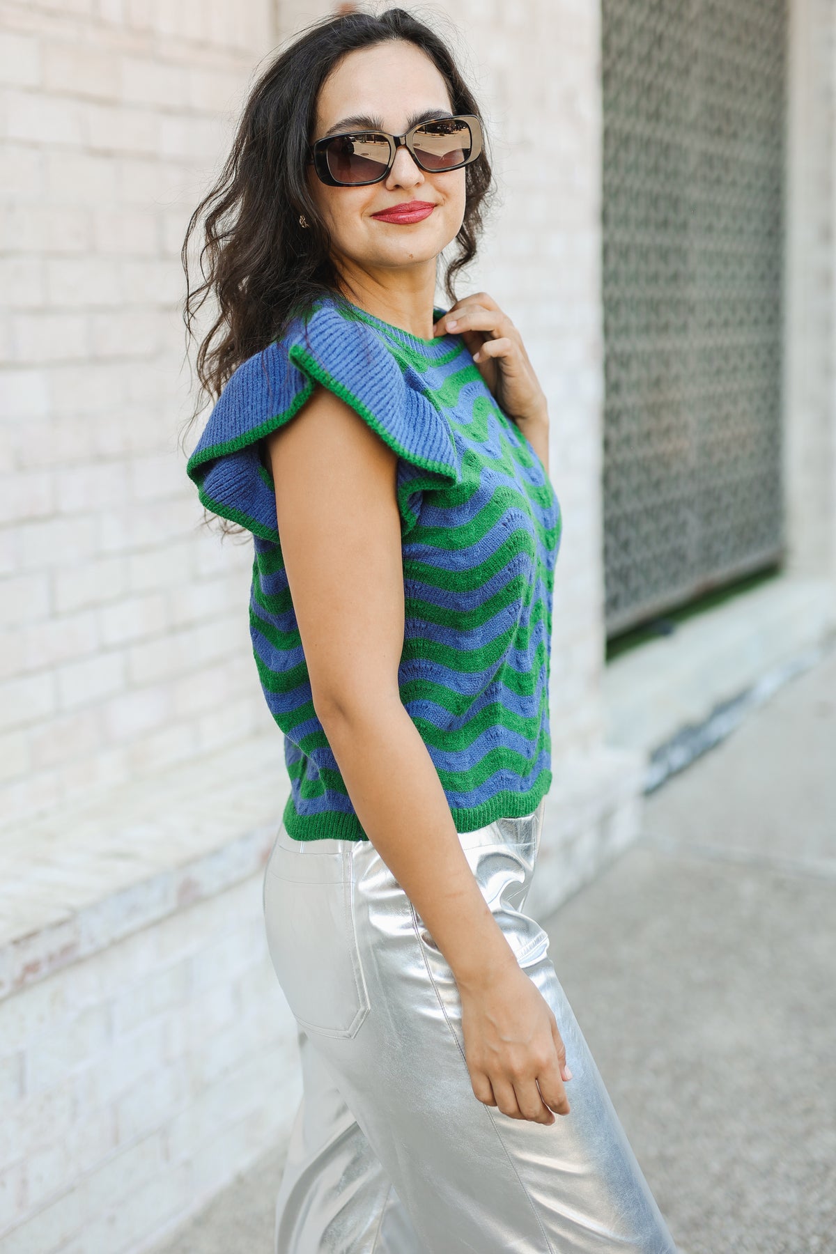 Woman wearing a blue and green patterned sleeveless top and silver pants against a light-colored wall.