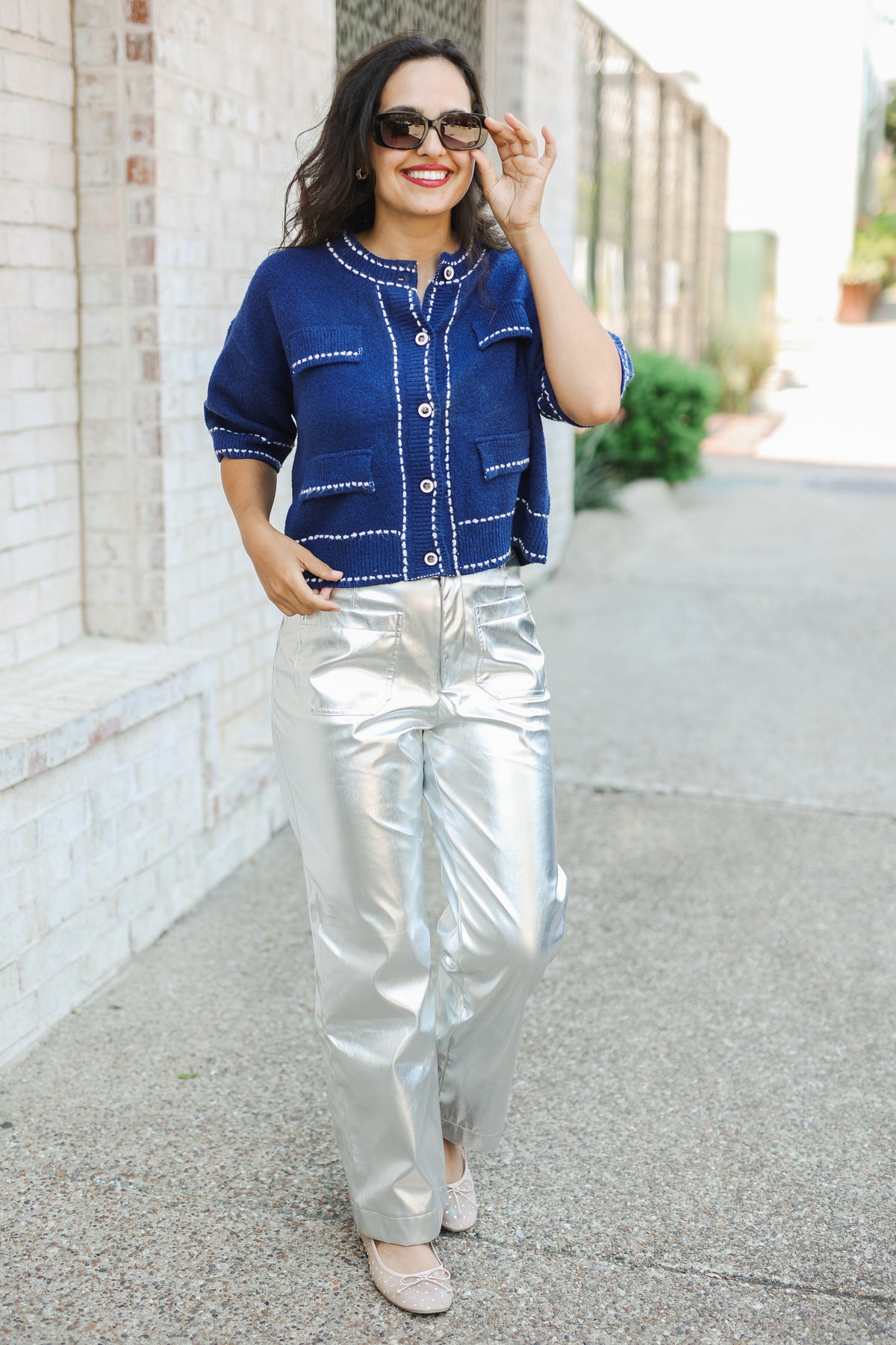 Woman wearing a blue top and silver pants standing on a sidewalk.