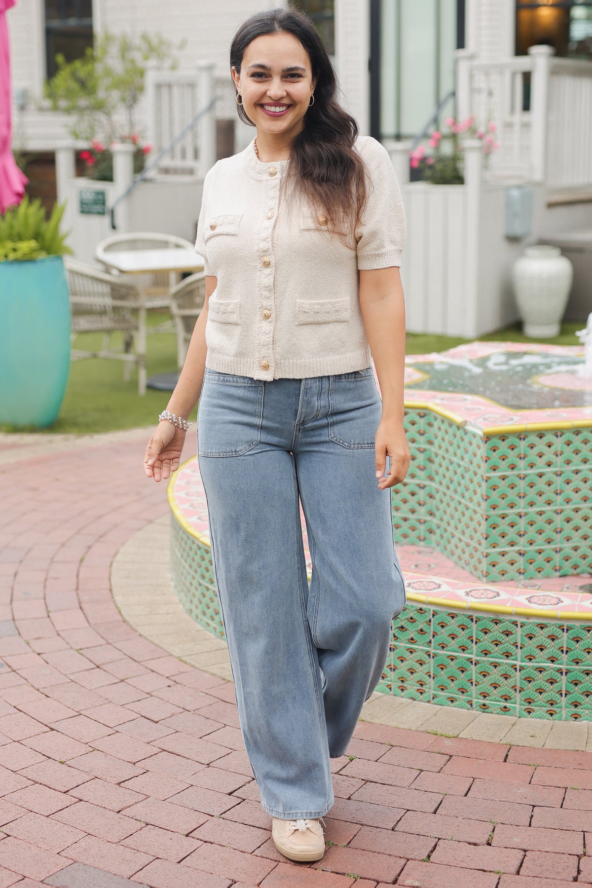 Woman in a beige cardigan and blue jeans standing in a garden area with plants and a small pond.