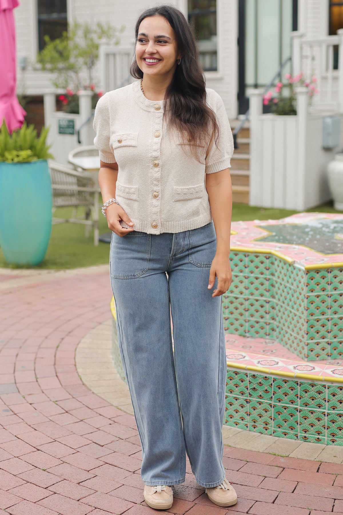 Woman in a light-colored cardigan and blue jeans standing on a patio with a garden in the background.