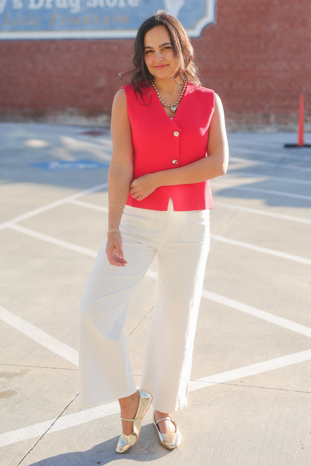 Woman wearing a red sleeveless top and white pants standing in a parking lot.