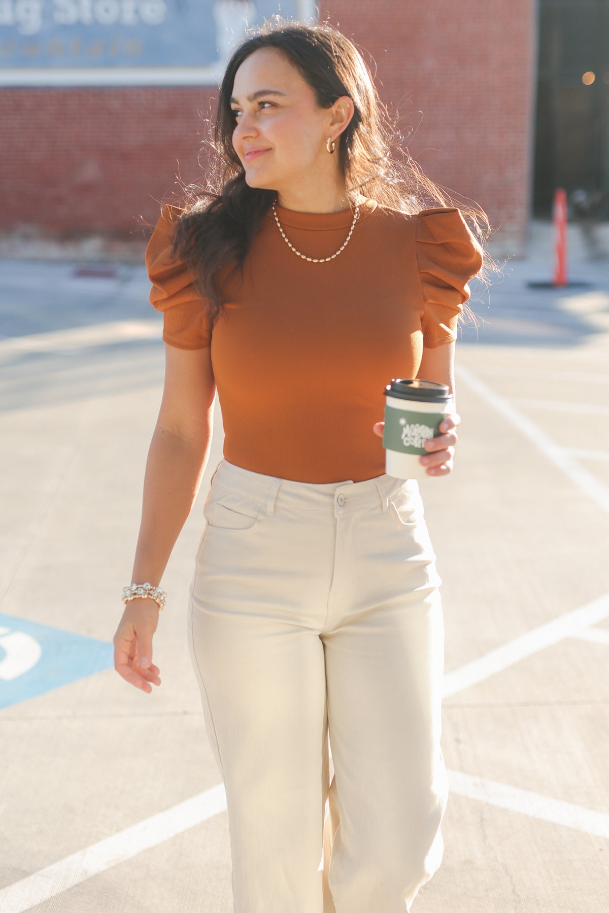 Woman in a rust-colored top and white pants holding a coffee cup outdoors.