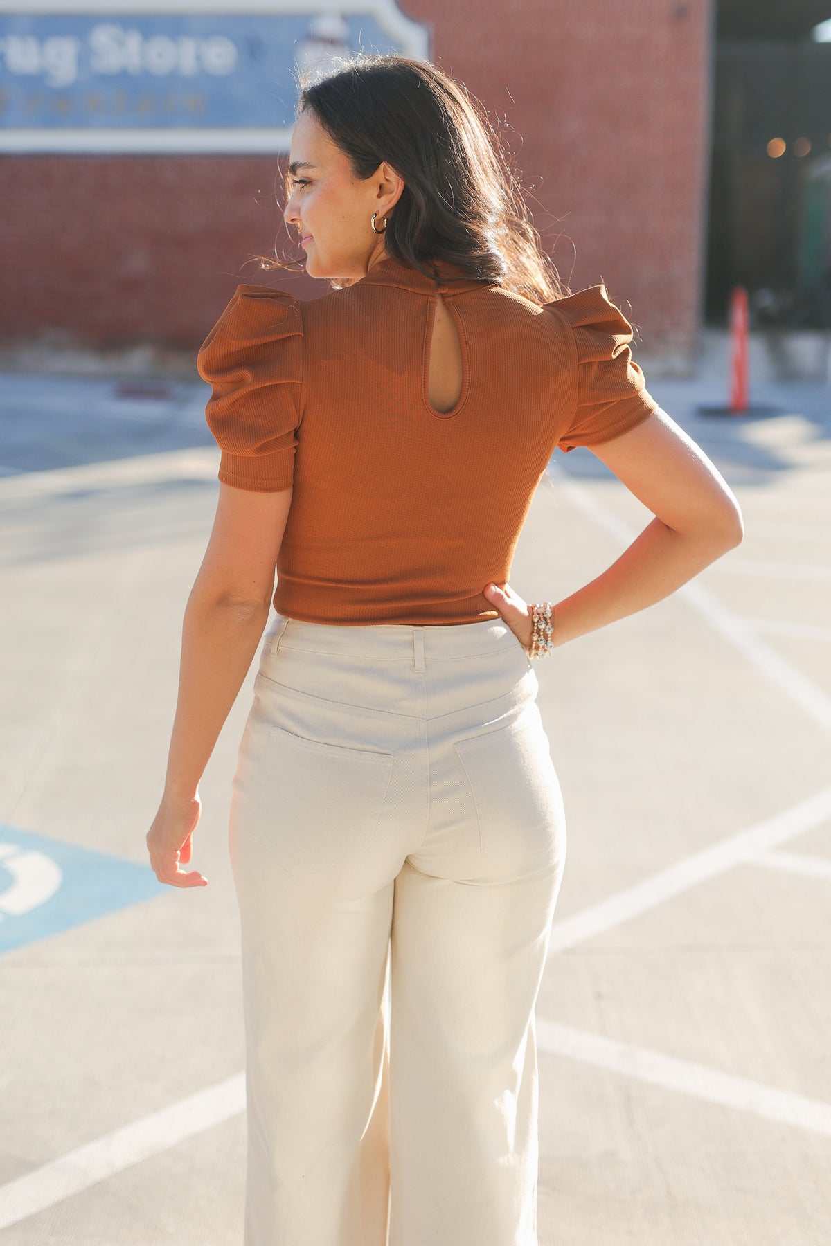 Woman wearing a brown top and white pants standing in a parking lot.