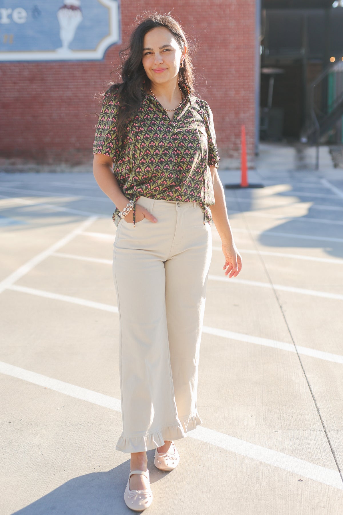 Woman standing in a parking lot wearing a patterned blouse and beige pants.