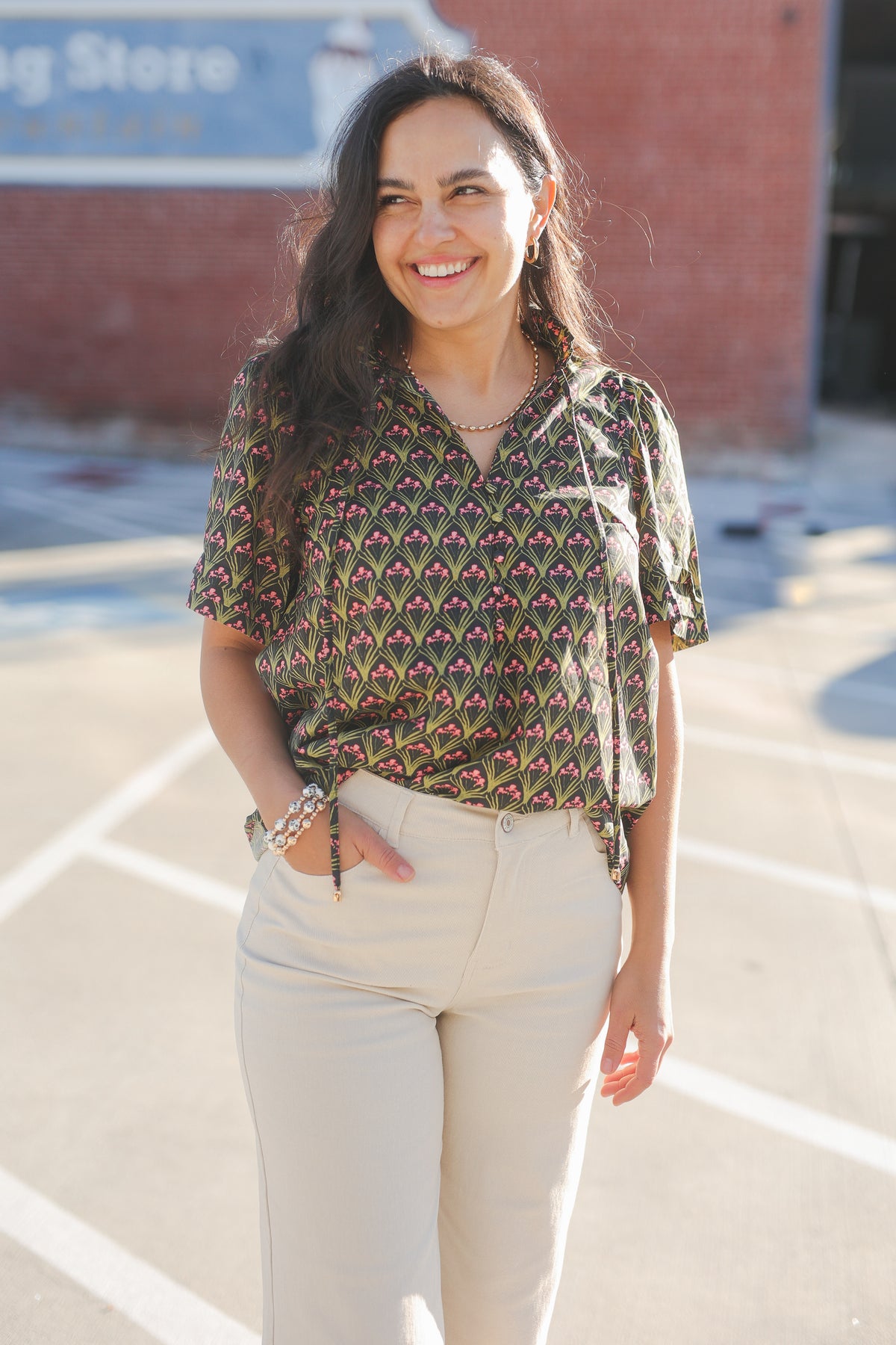 Woman wearing a patterned blouse and beige pants standing outdoors.