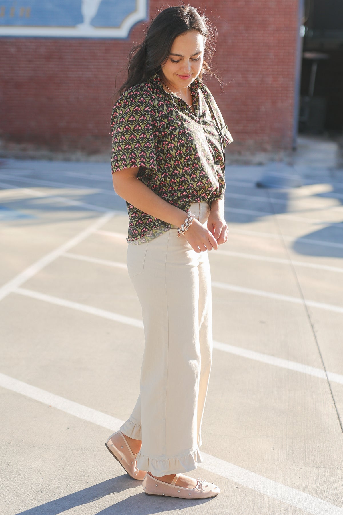 Woman standing in a parking lot wearing a patterned top and white pants.