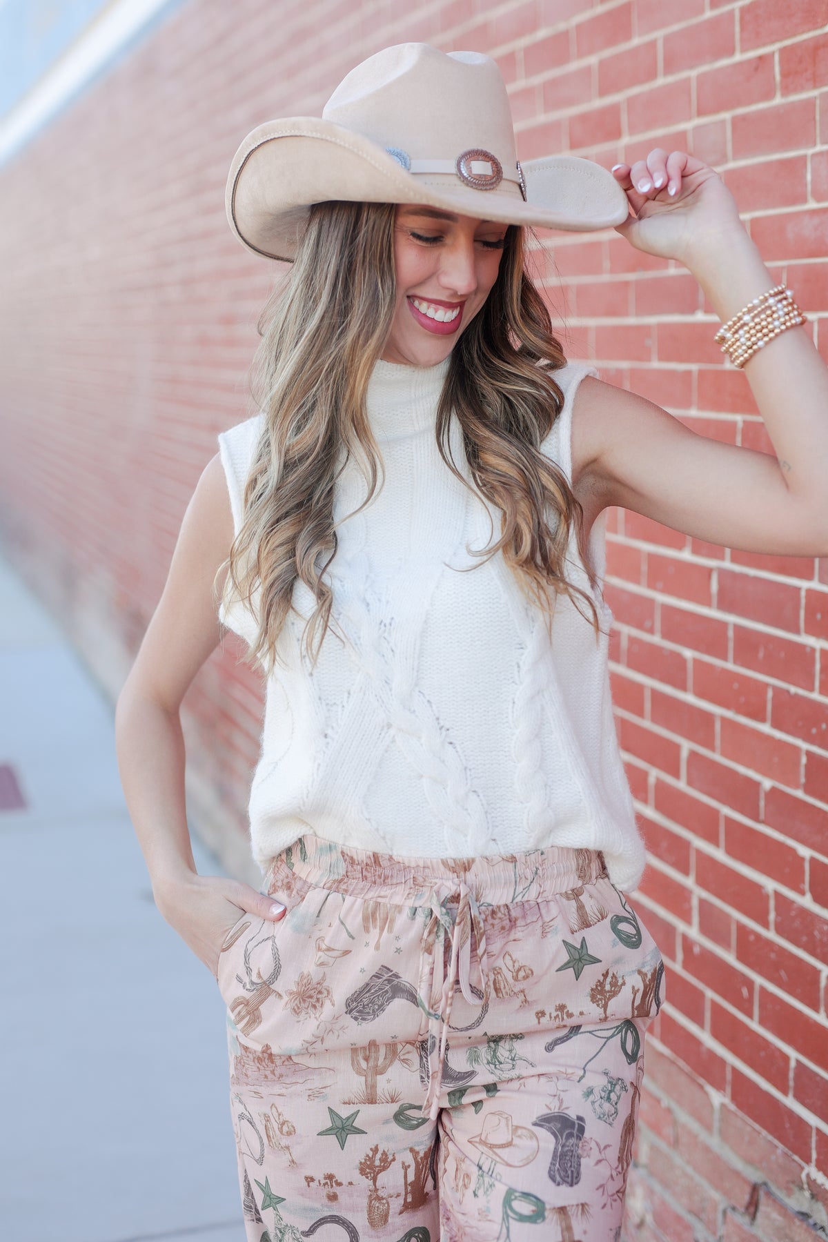 Woman wearing a white sleeveless top and patterned pants against a brick wall.