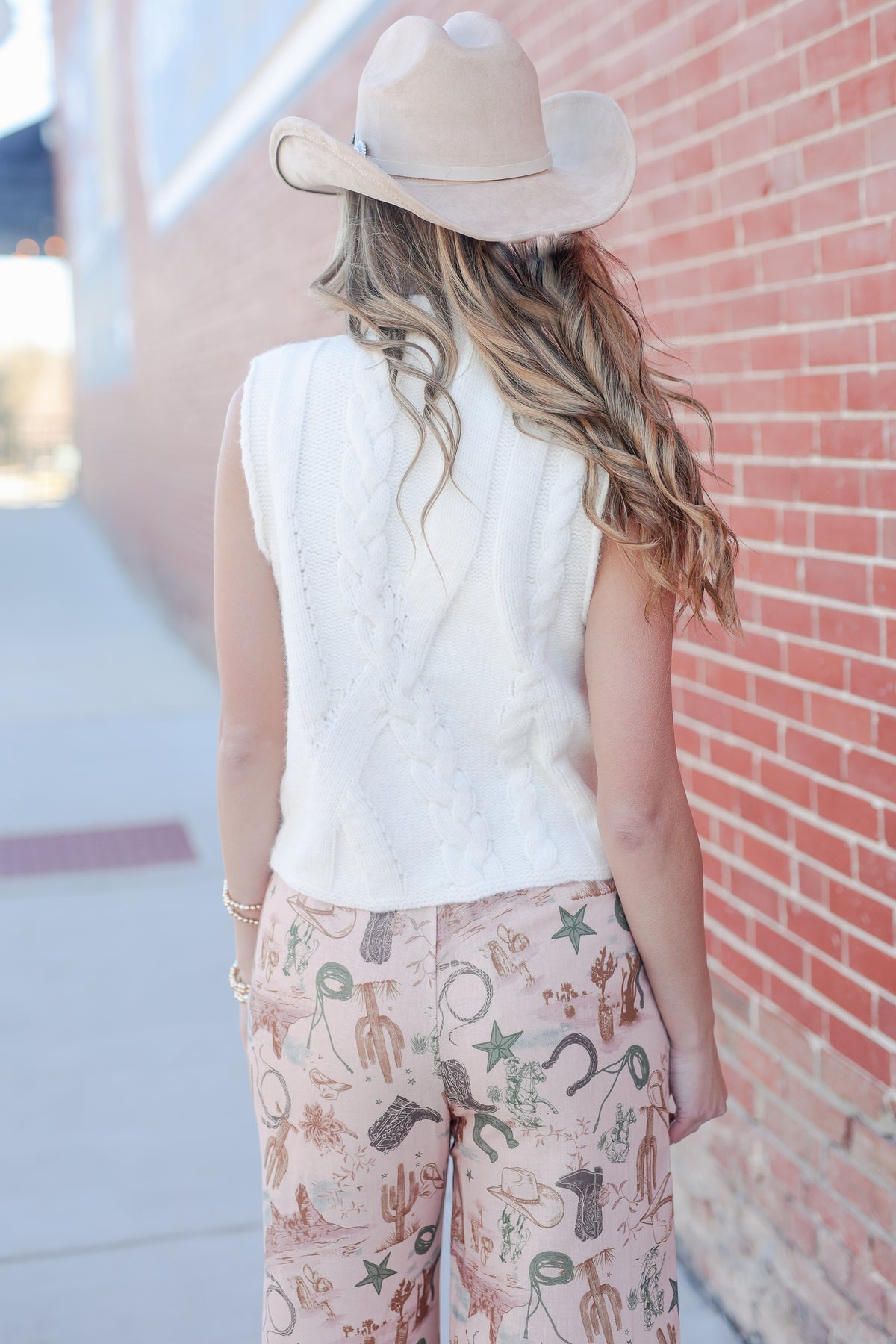 Person wearing a white sleeveless top and patterned pants with a cowboy hat, standing against a brick wall.