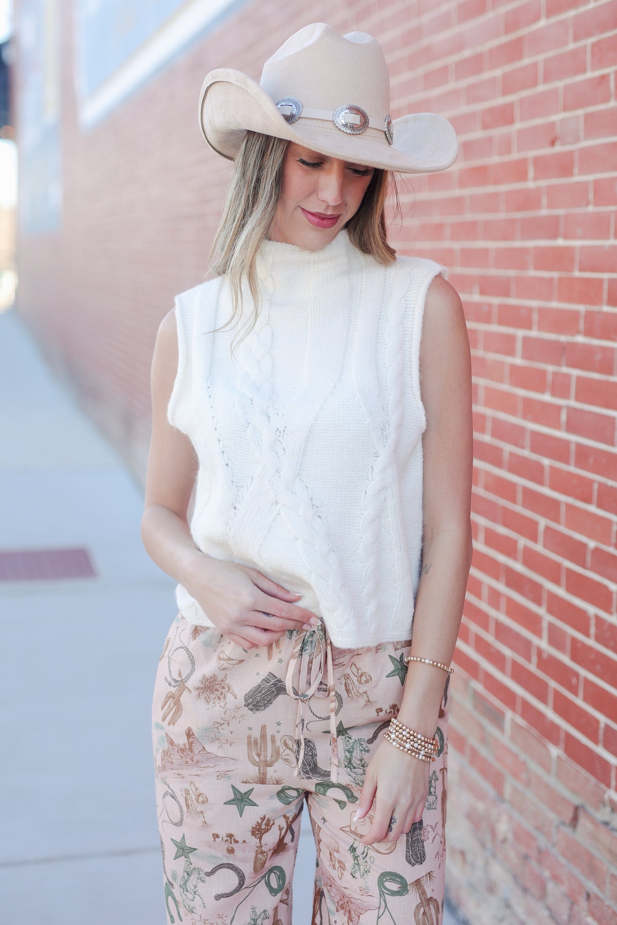 Woman wearing a white sleeveless top and patterned pants with a beige cowboy hat against a brick wall.