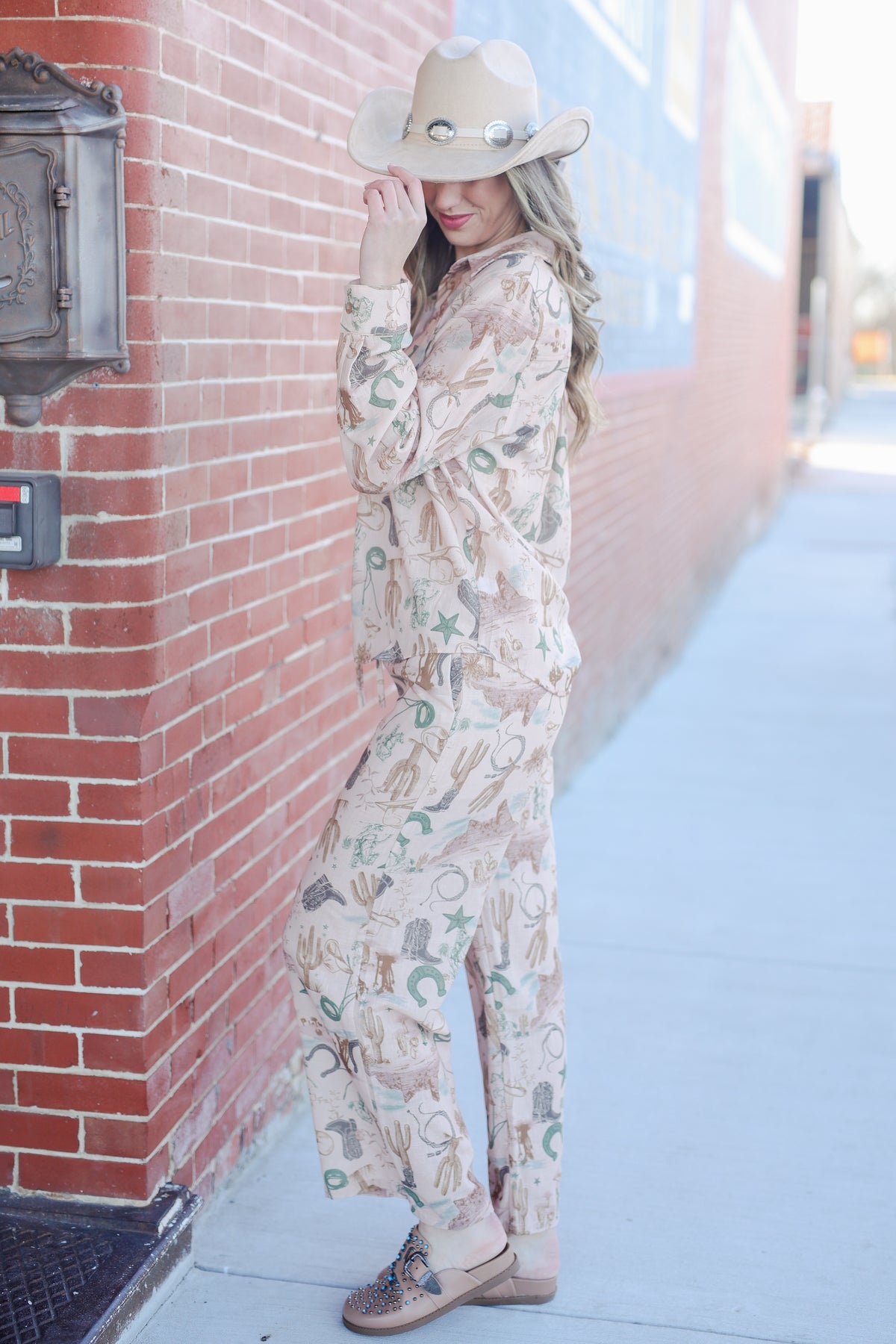 Woman in a patterned outfit and hat leaning against a brick wall.