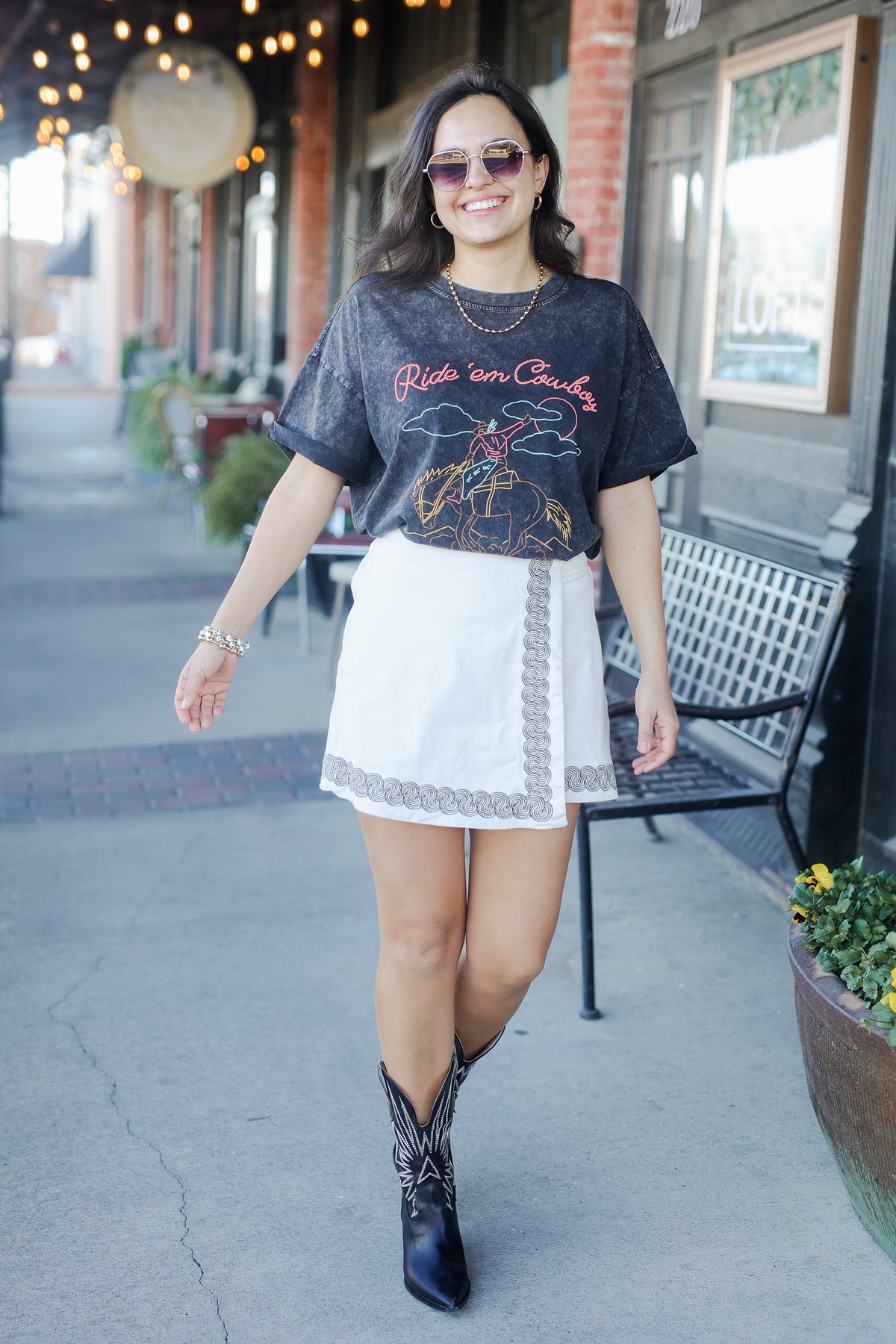 Woman wearing a graphic t-shirt, white skirt, and cowboy boots on a sidewalk.