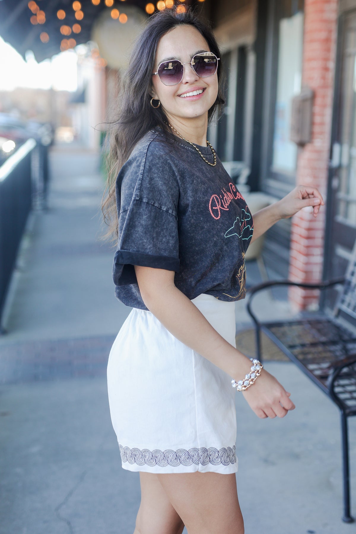 Woman wearing sunglasses and a dark gray t-shirt with 'Rice' printed on it, standing on a sidewalk.