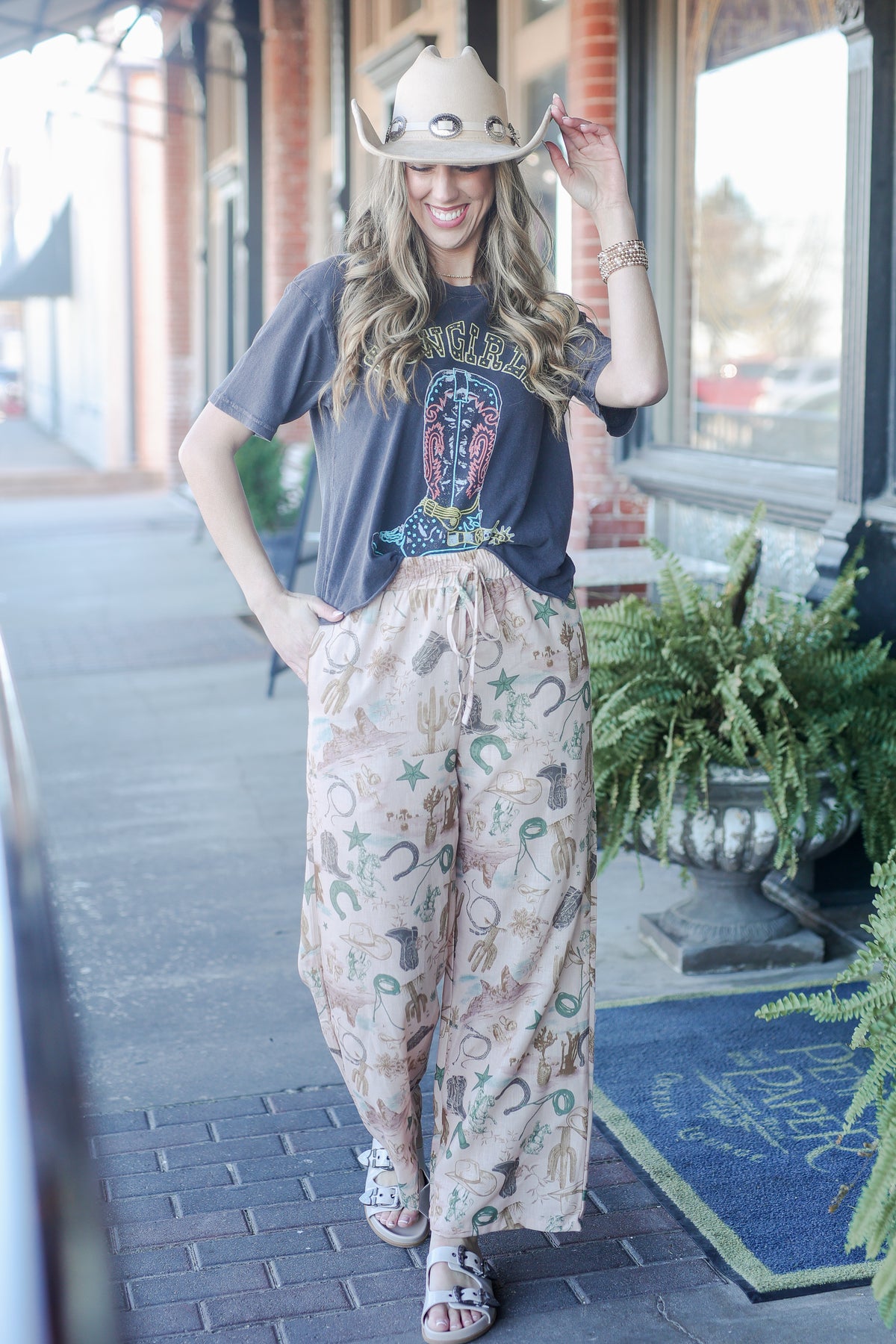 Woman wearing a cowboy hat and graphic t-shirt with patterned pants on a sidewalk.