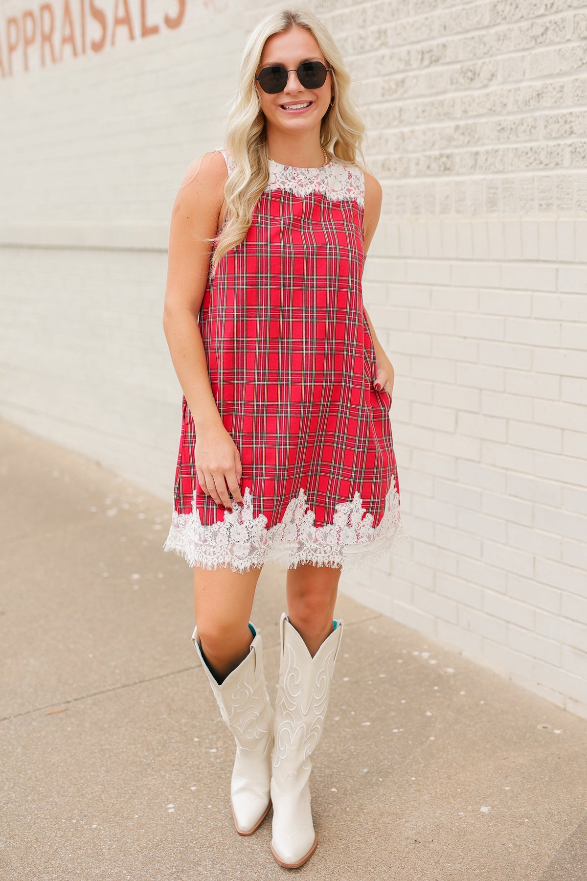 Blonde model standing outside wearing the Hollybrook Plaid Lace Mini Dress in red, green, and black tartan plaid featuring an ivory floral lace neckline and lace-trimmed hem with the white Senora boots from Southern House Clothing