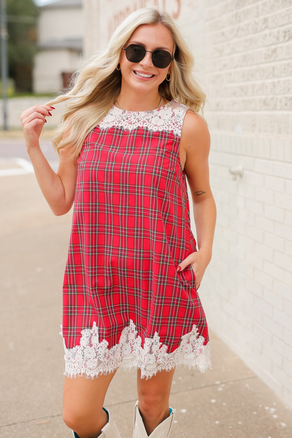 Blonde model standing outside wearing the Hollybrook Plaid Lace Mini Dress in red, green, and black tartan plaid featuring an ivory floral lace neckline and lace-trimmed hem with the white Senora boots from Southern House Clothing