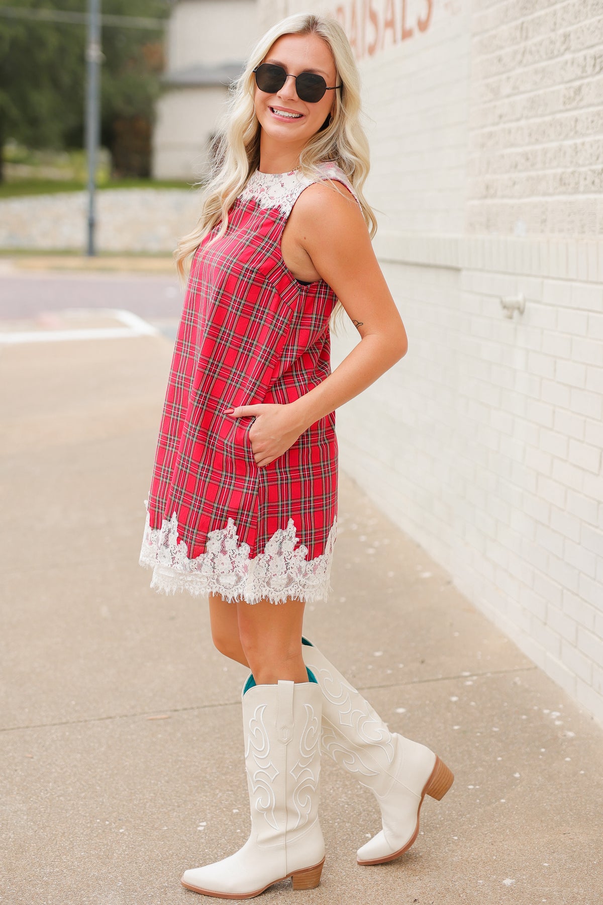 Blonde model standing outside wearing the Hollybrook Plaid Lace Mini Dress in red, green, and black tartan plaid featuring an ivory floral lace neckline and lace-trimmed hem with the white Senora boots from Southern House Clothing