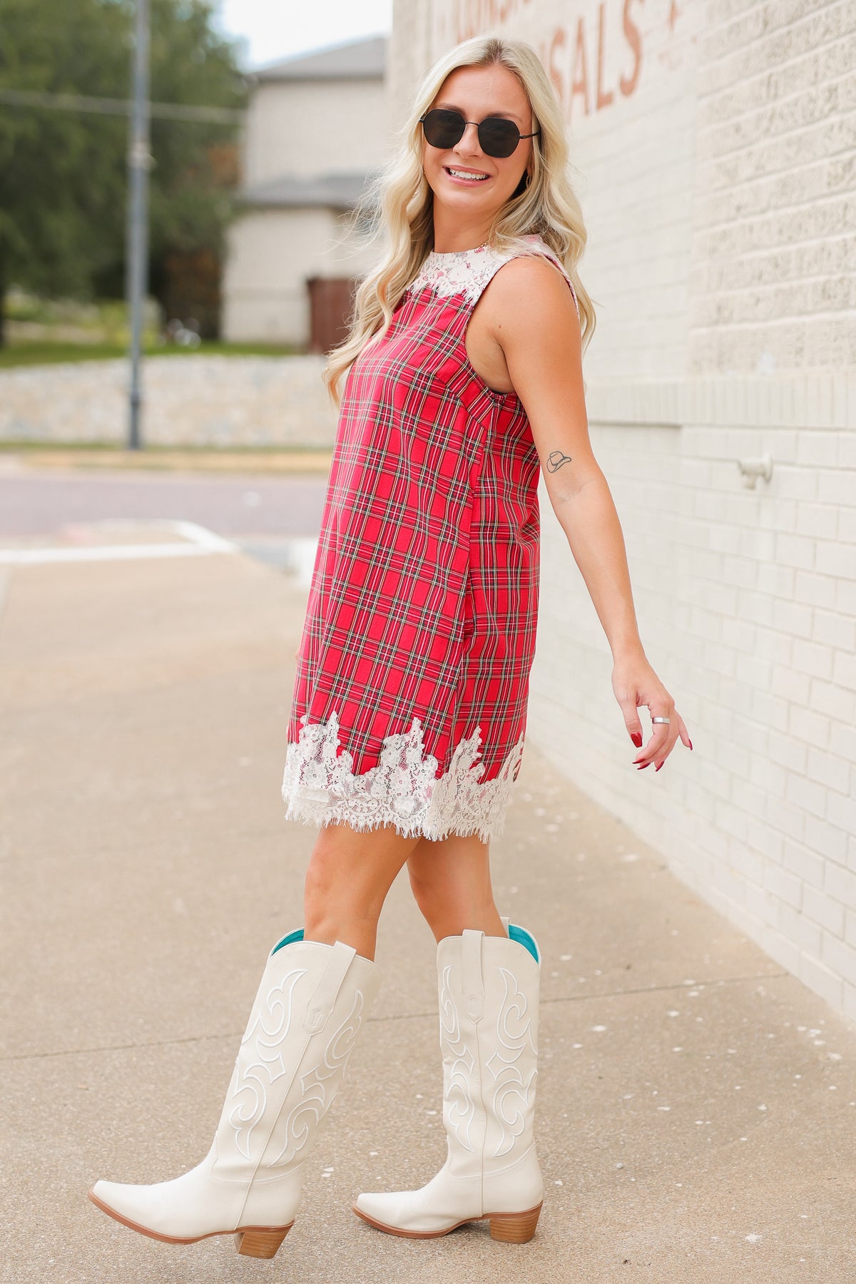Blonde model standing outside wearing the Hollybrook Plaid Lace Mini Dress in red, green, and black tartan plaid featuring an ivory floral lace neckline and lace-trimmed hem with the white Senora boots from Southern House Clothing
