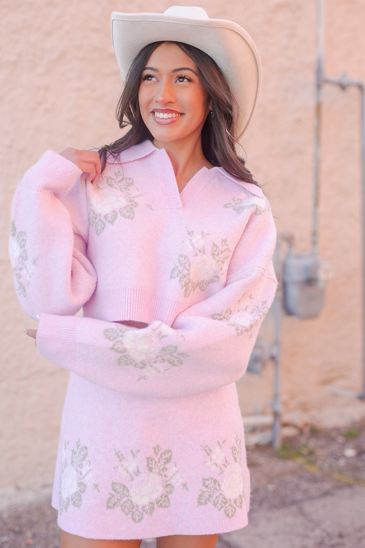 Woman wearing a pink embroidered dress and cowboy hat against a beige wall.