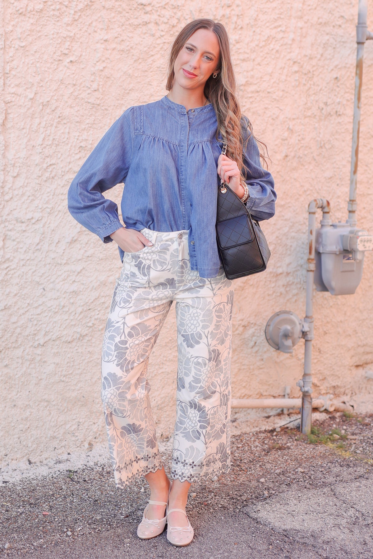 Woman wearing a blue blouse and floral pants standing against a beige wall.