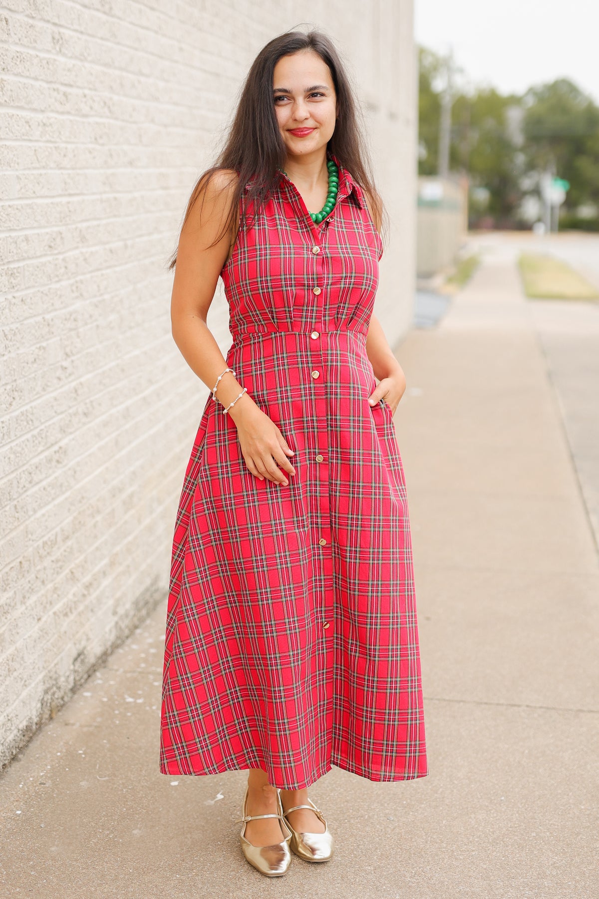 Model wearing the Mistletoe Manor Plaid Maxi Dress in Red Multi, featuring a sleeveless collared neckline, gold-tone button-front detailing, defined waist, and flowing A-line maxi skirt styled with metallic gold ballet flats standing outside. Southern House Clothing