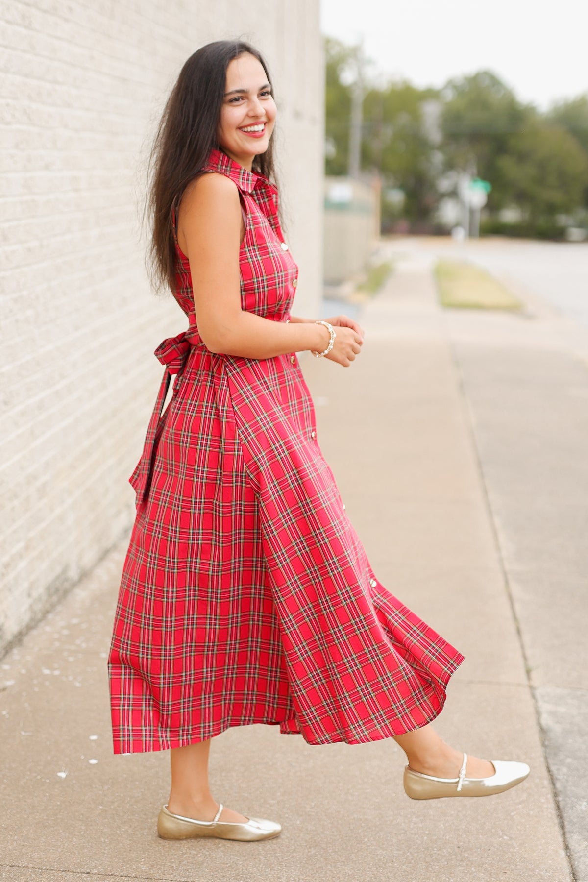 Model wearing the Mistletoe Manor Plaid Maxi Dress in Red Multi, featuring a sleeveless collared neckline, gold-tone button-front detailing, defined waist, and flowing A-line maxi skirt styled with metallic gold ballet flats standing outside. Southern House Clothing